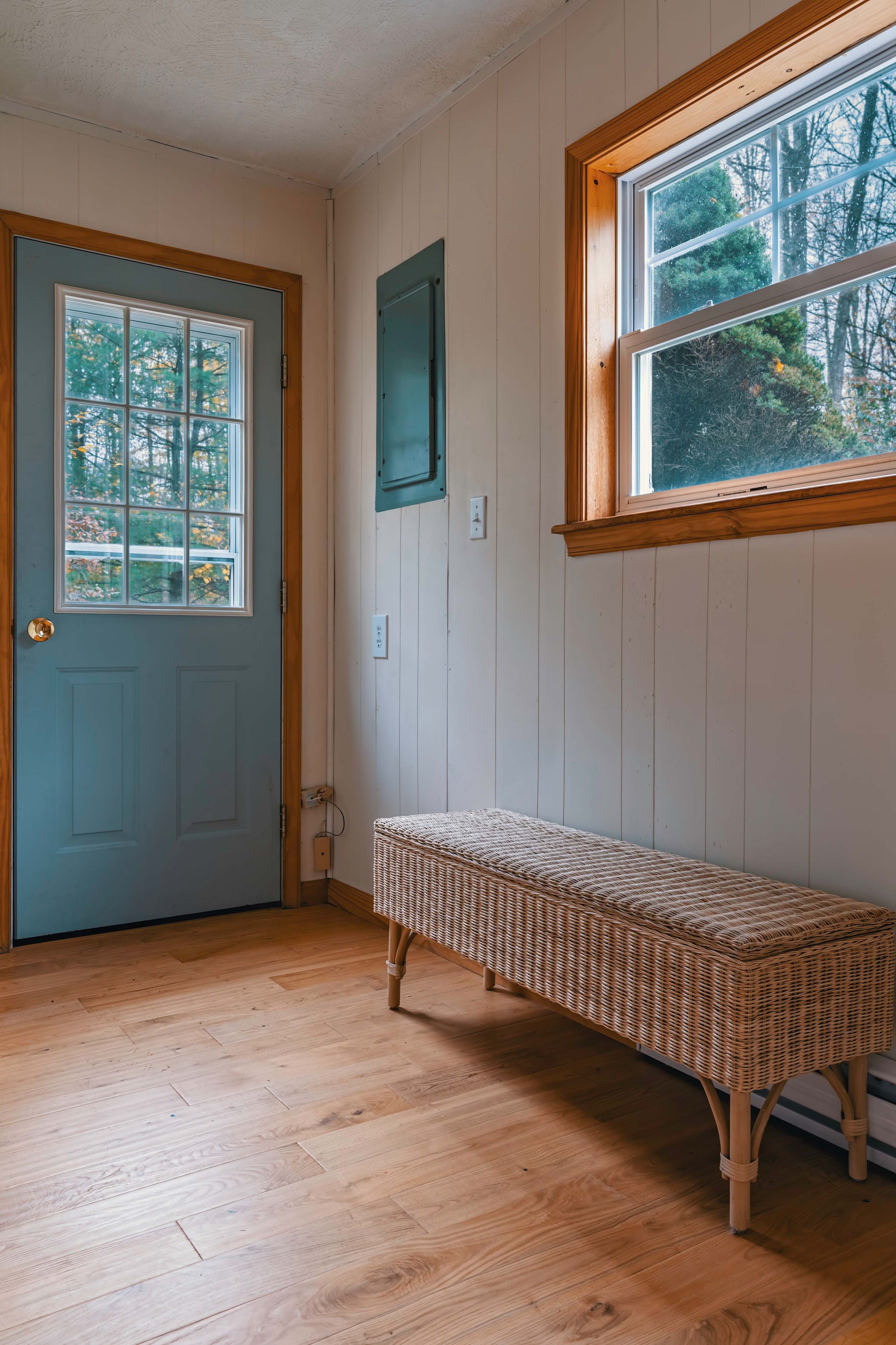 Simple entryway with light wood floors and white paneled walls, featuring a light blue door with glass panes leading outside. A woven bench sits beneath a window, providing a functional and decorative element.