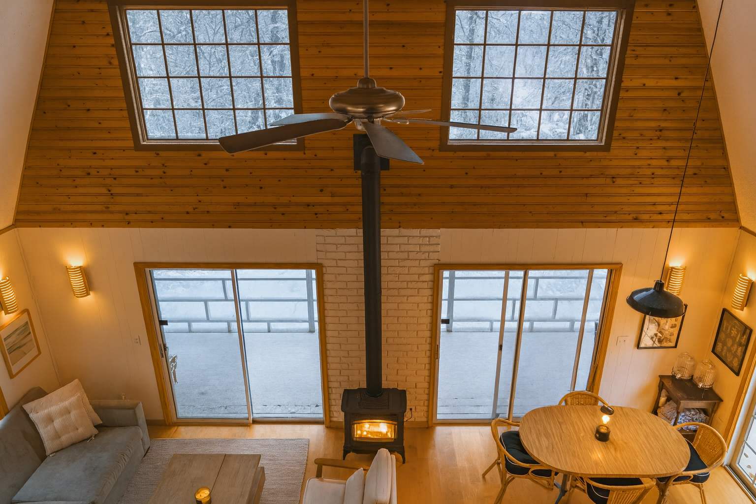 High-angle view of a cozy living space with a central wood-burning stove, vaulted wood-paneled ceiling, and large grid windows overlooking a snowy landscape, creating a warm, inviting atmosphere.