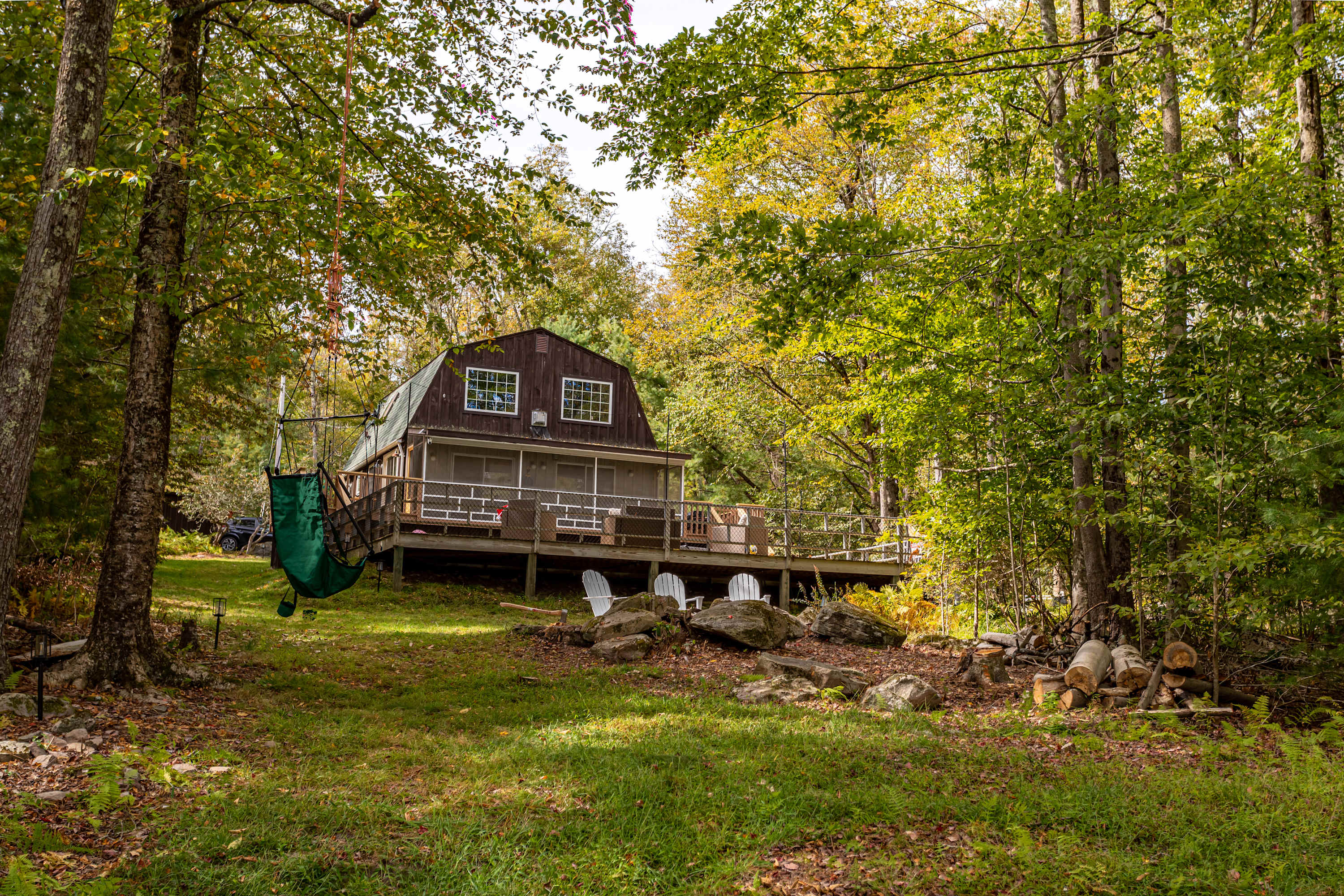 View of the back of the dark brown barn-style house with a screened porch and a large wooden deck, nestled among tall green and yellow trees. A green hammock chair hangs from a tree, and white Adirondack chairs are arranged around a fire pit area in the grassy yard.