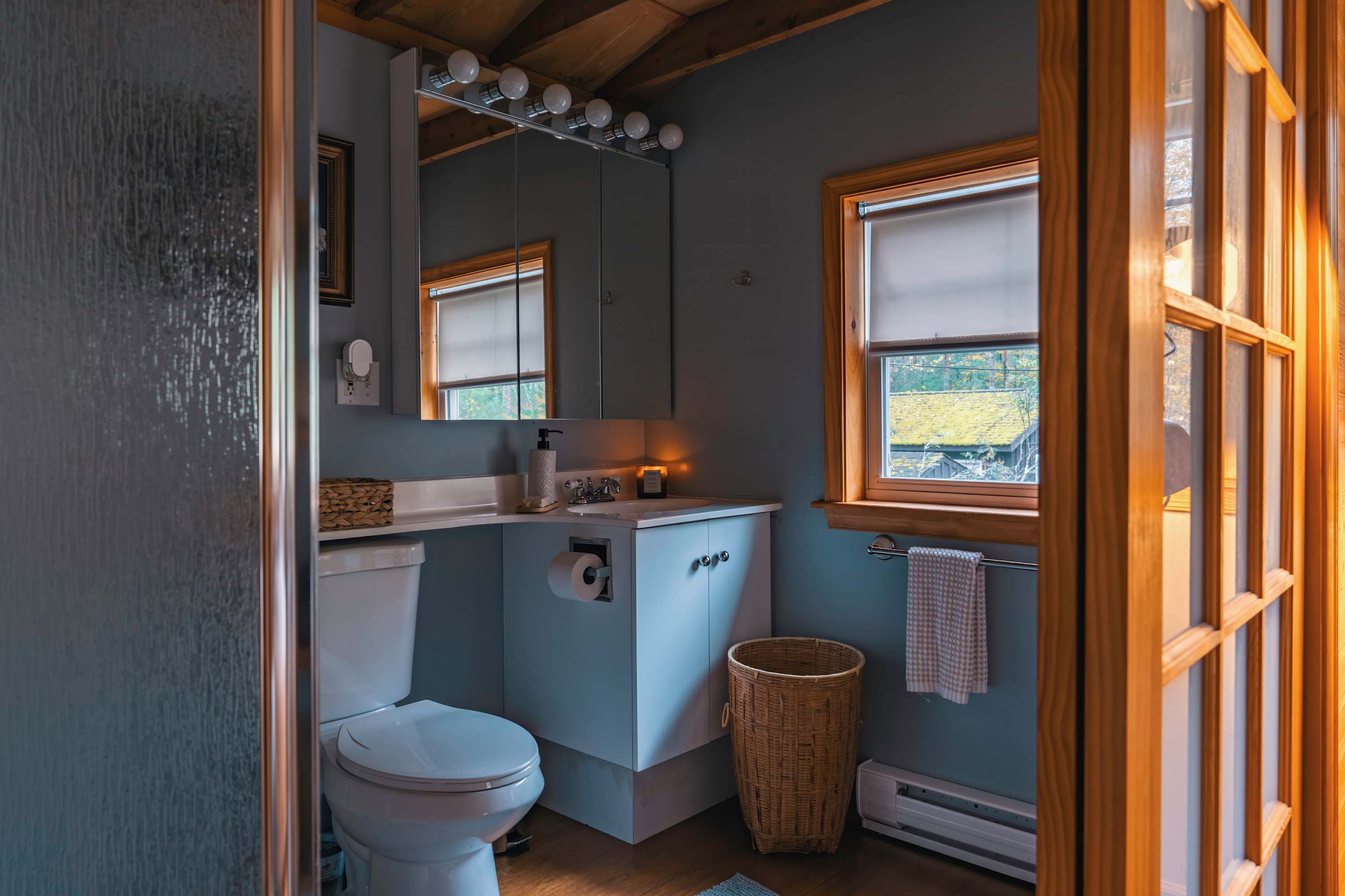 Compact bathroom with a white toilet, a vanity with a medicine cabinet and mirror, and a window, featuring light blue walls and warm wood trim, with a wicker basket for storage.