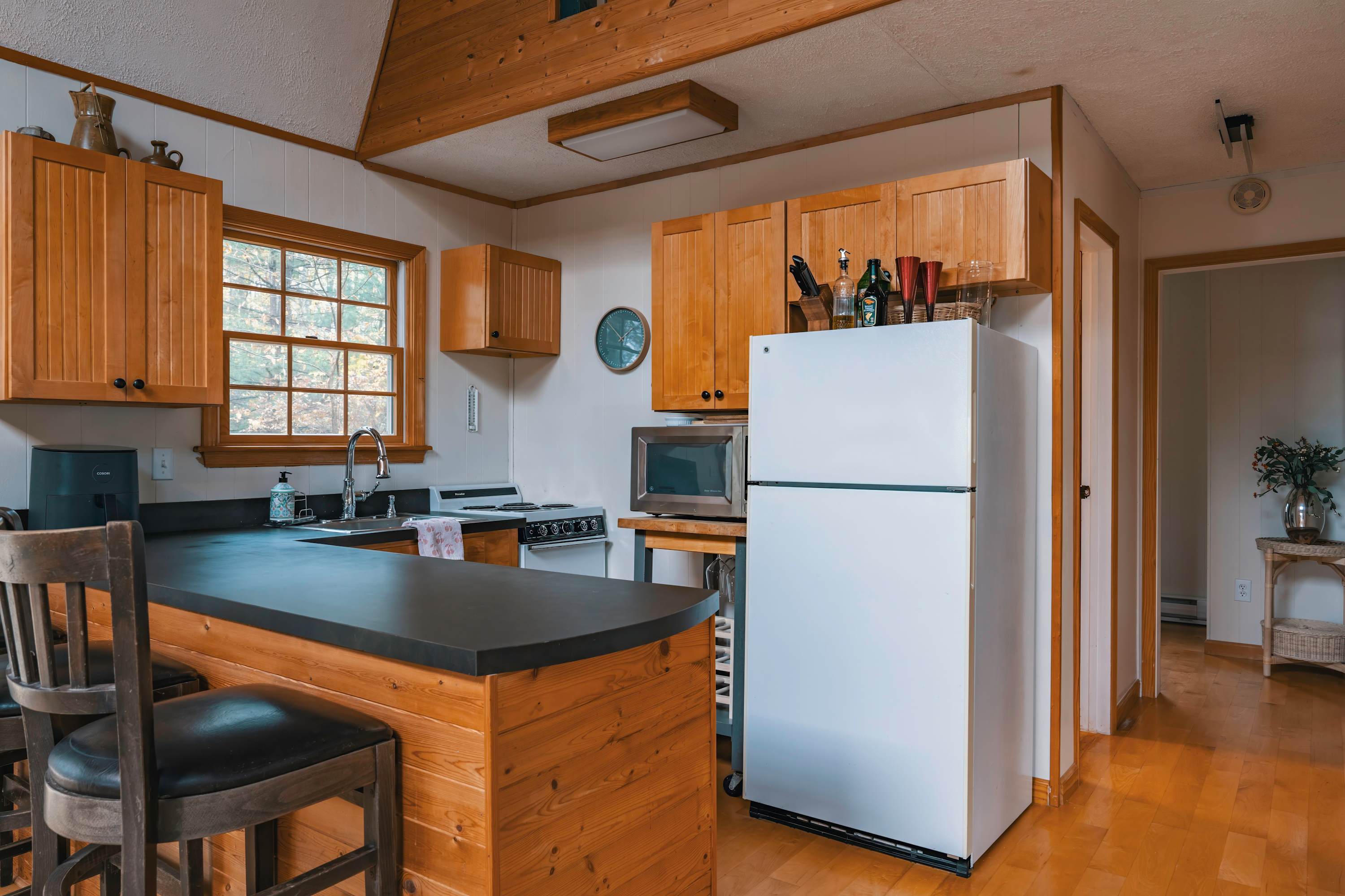 Well-equipped kitchen featuring light wood cabinets, a black countertop, a stainless steel sink, and a white stove, with a window overlooking autumn trees and a bar area with stools.