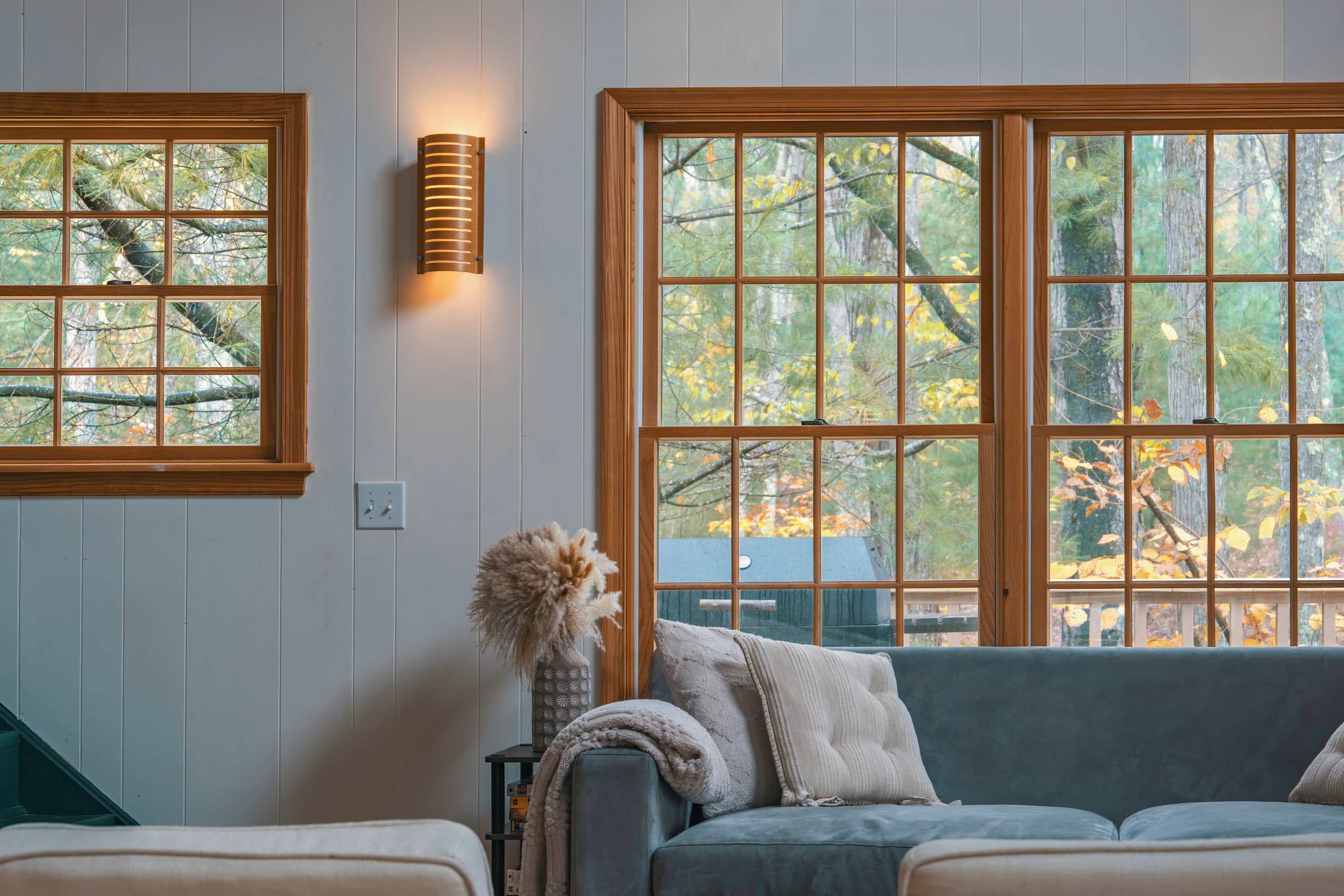 Close-up of a living room corner with a light blue sofa adorned with decorative pillows and a throw blanket. Large windows with wooden frames offer a view of the outdoor greenery, and a decorative vase with dried pampas grass sits on a side table.