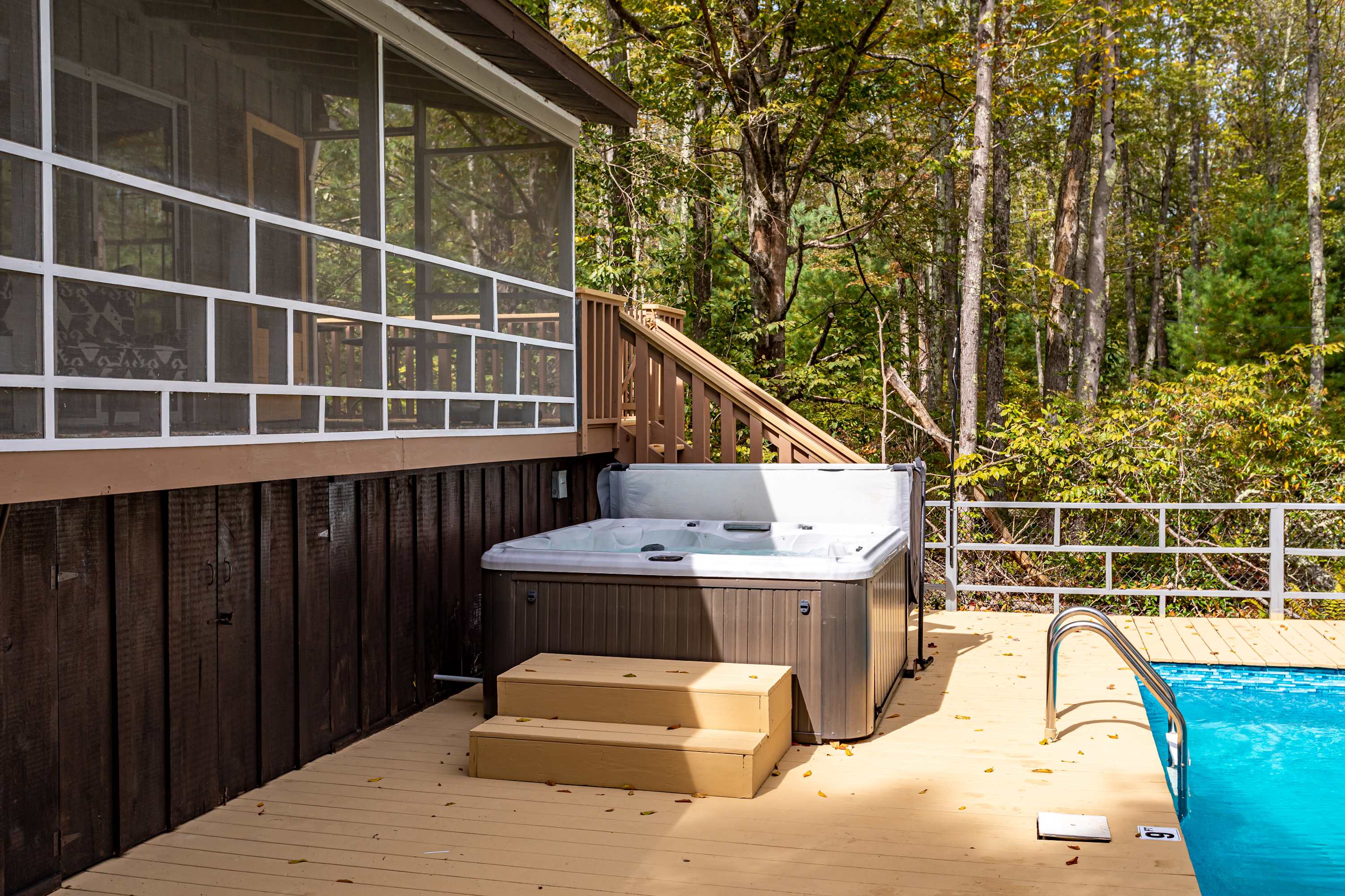 A modern hot tub with wooden steps sits on a light-colored wooden deck, adjacent to a screened porch and a swimming pool. The area is surrounded by tall trees, creating a private outdoor oasis.