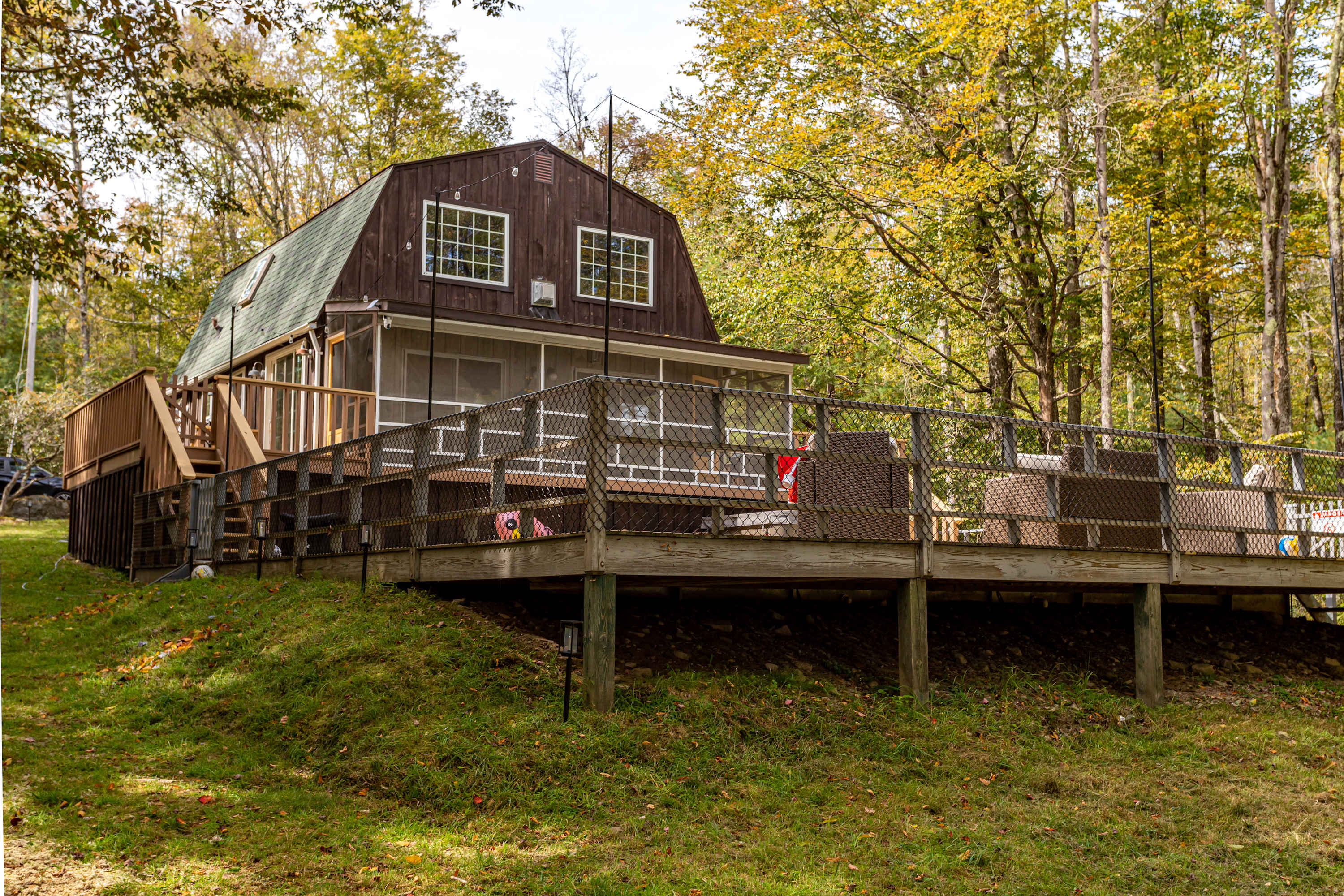 Exterior view of a brown barn-style house nestled among lush trees, featuring a large wooden deck with a metal railing. The deck extends around the side of the house, offering ample outdoor living space on a gentle slope.