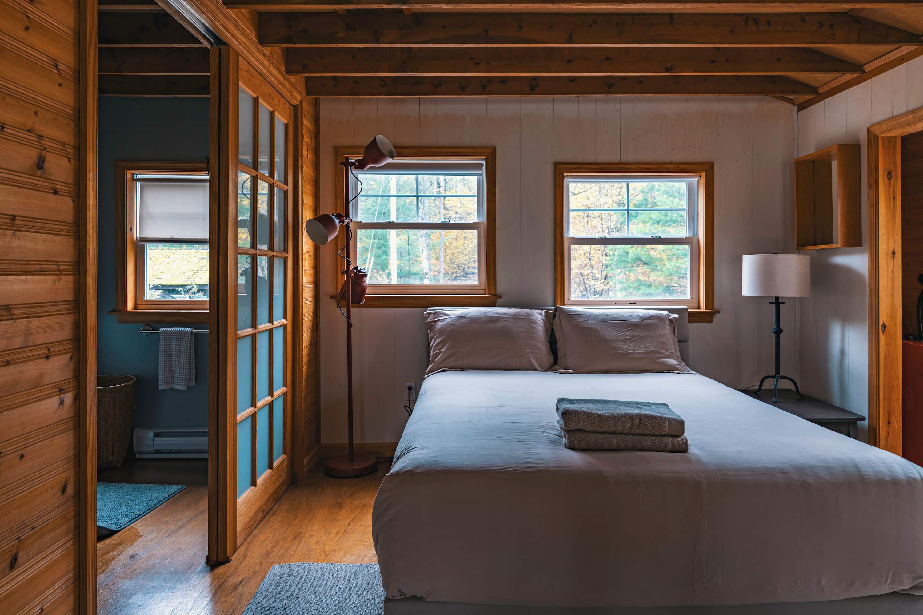 Cozy bedroom featuring a queen-sized bed with light bedding, two large windows overlooking autumn foliage, and warm wood paneling on walls and ceiling beams, with a partial view into an adjacent bathroom.