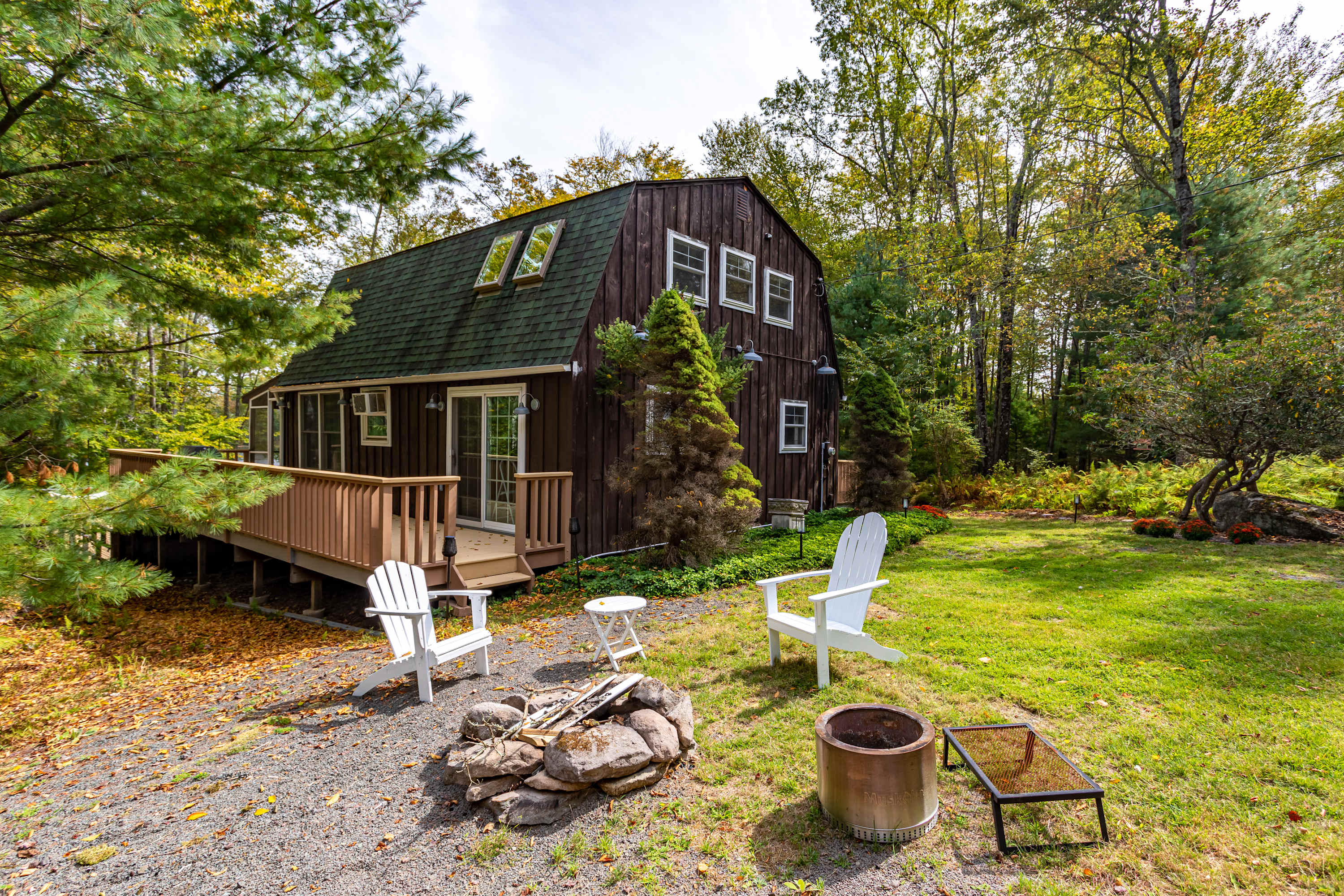 Front/side view of the dark brown barn-style house with a green roof and multiple windows, surrounded by a well-maintained lawn and tall trees. A gravel area with a stone fire pit and white Adirondack chairs is in the foreground.