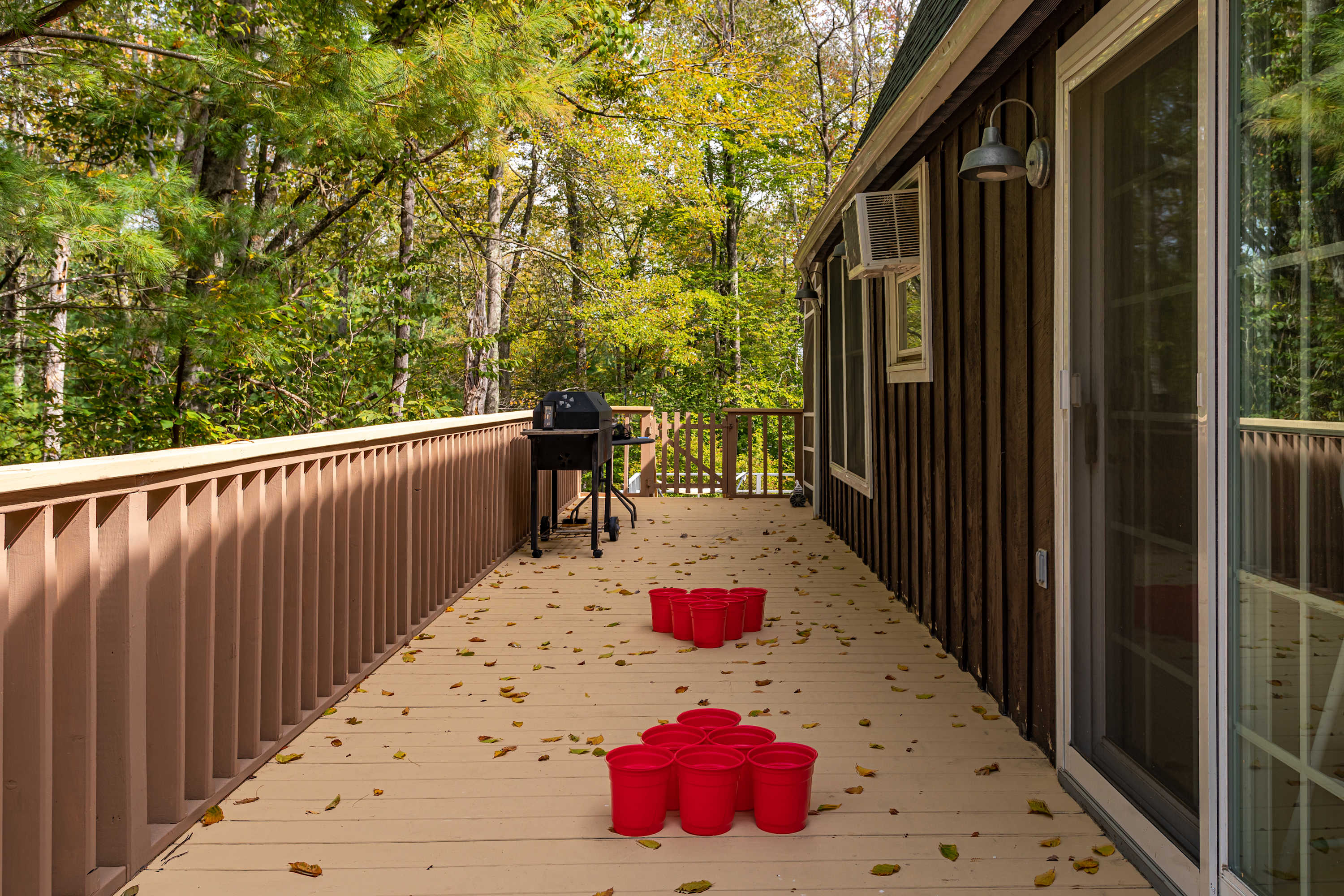 A long, light-colored wooden deck with a brown railing features a black BBQ grill and several red buckets, with fallen leaves scattered across the surface, next to a sliding glass door of the house.