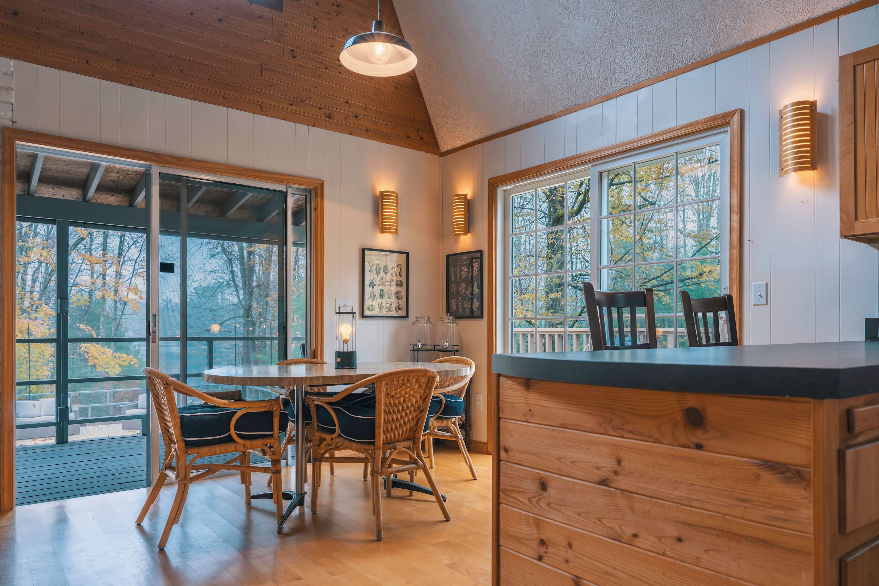 Bright dining area with a round wooden table and four wicker chairs, situated next to large sliding glass doors leading to a screened porch, with a view of the kitchen bar area.