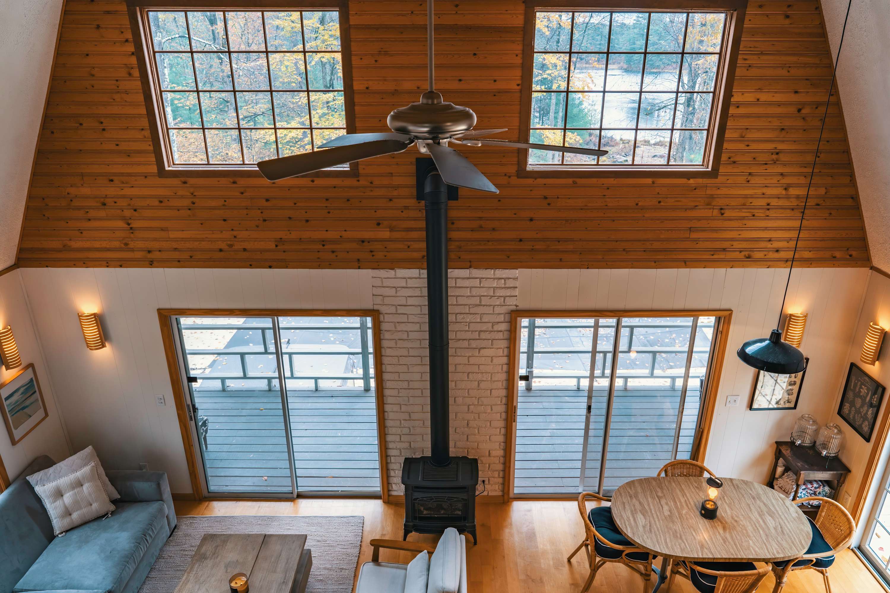Overhead view of the living and dining area, showcasing the high vaulted ceiling with exposed wooden beams and a central ceiling fan. Large windows overlook the lake, and a black wood-burning stove anchors the center of the room, surrounded by seating and a dining table.