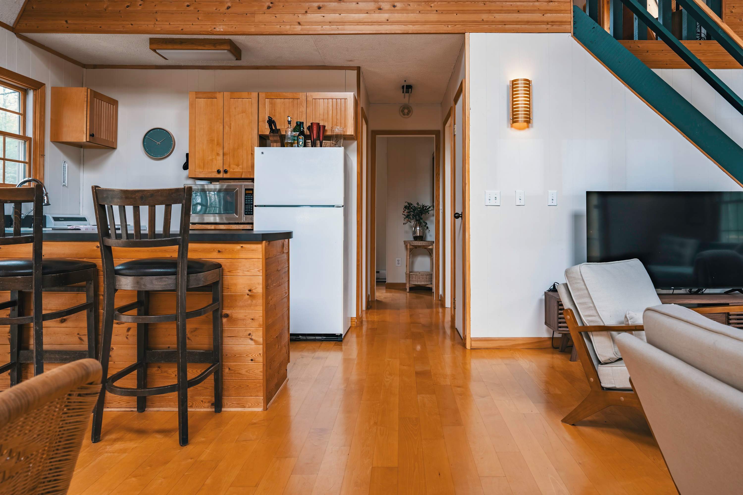 Open-concept kitchen with light wood cabinetry, a white refrigerator, and a black countertop island with bar stools, flowing into a living area with a sofa and TV, all on polished hardwood floors.