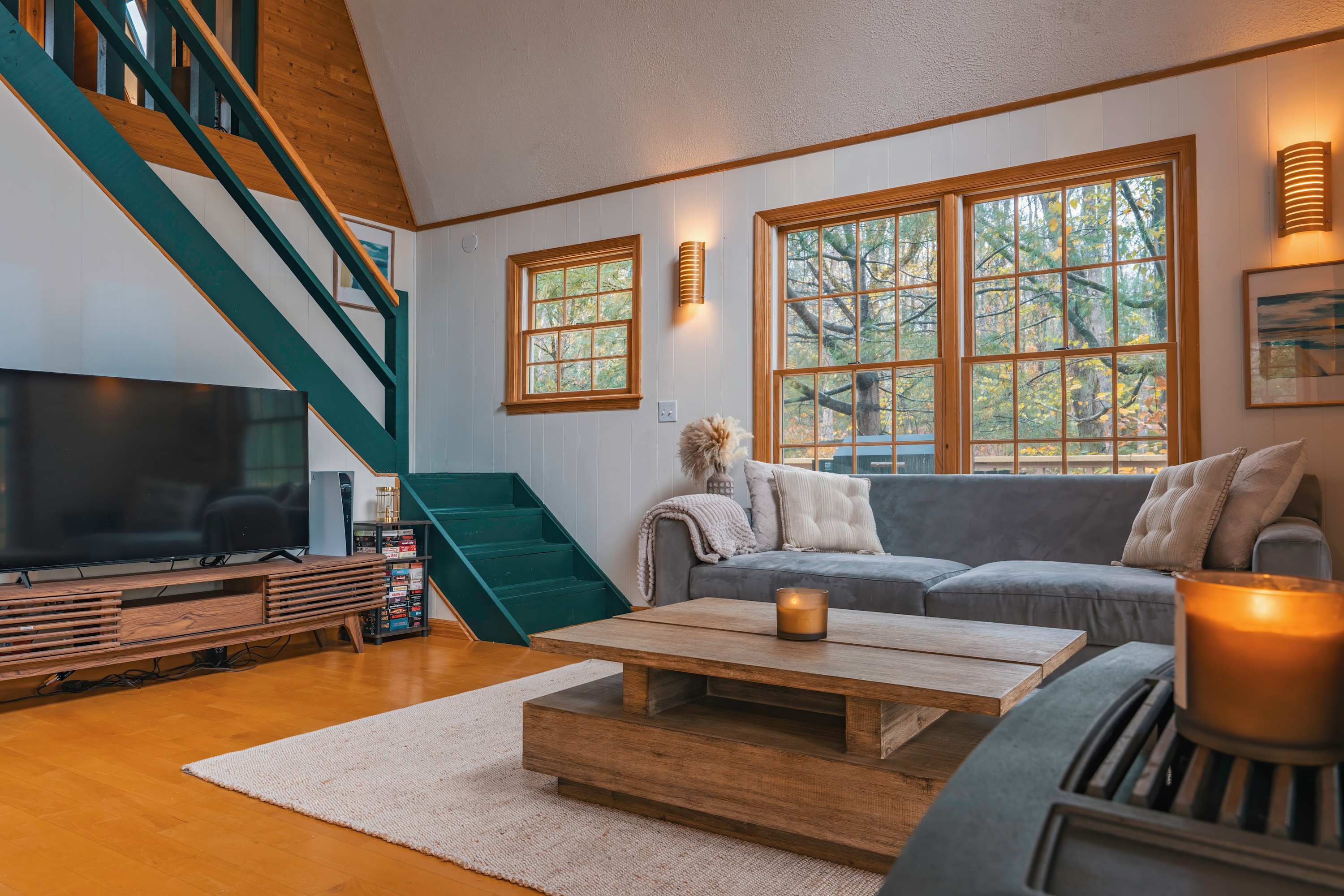 Cozy living room featuring a grey sectional sofa, a rustic wooden coffee table, a flat-screen TV on a console, and a unique green-painted staircase leading to an upper level, with large windows overlooking trees.