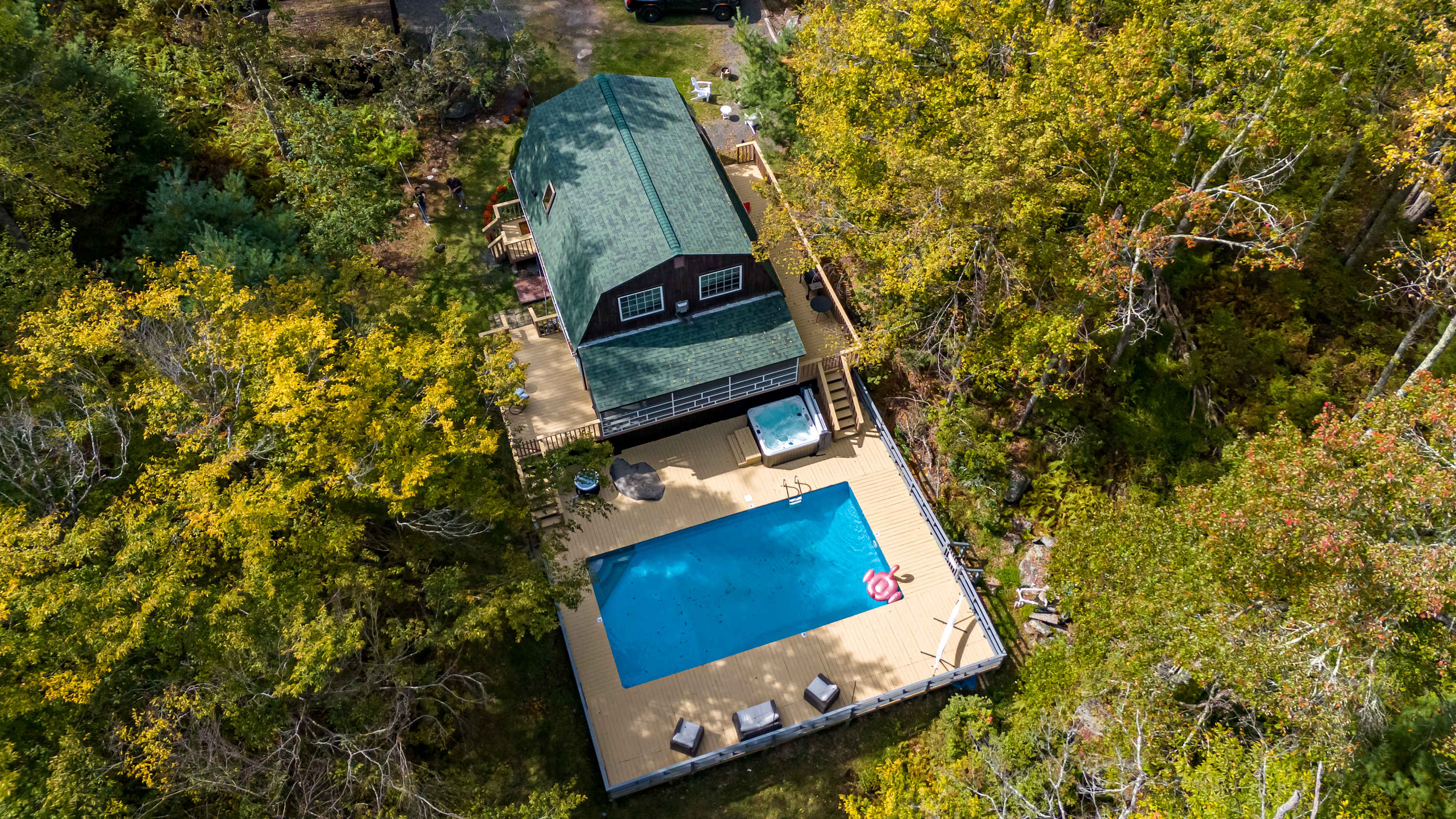 An overhead aerial shot reveals a long, dark-roofed house with a spacious wooden deck, featuring a bright blue swimming pool and a hot tub, completely enveloped by a lush, autumnal forest with yellow and green foliage.