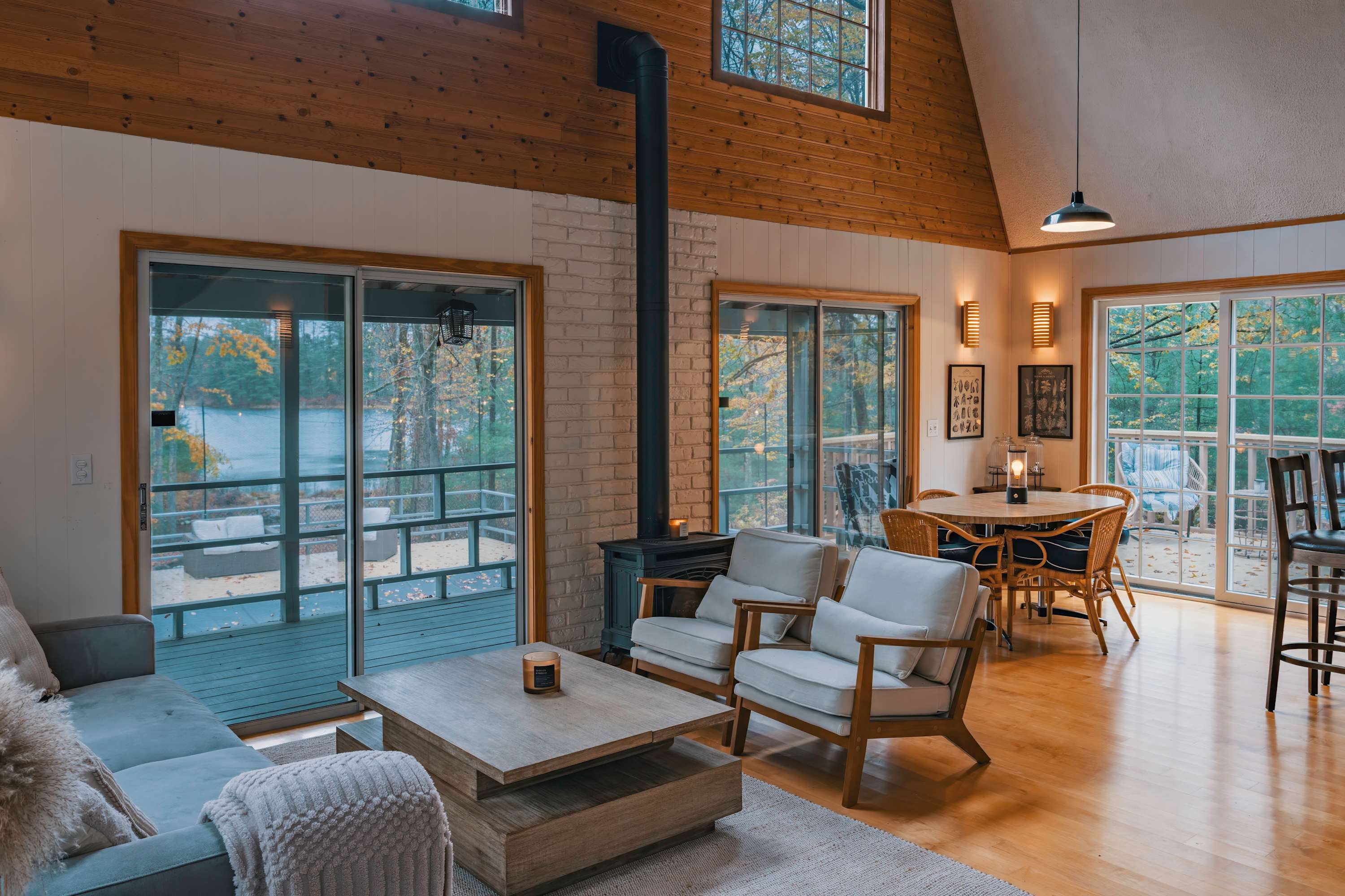 Open-concept living and dining area with a central wood-burning stove, light hardwood floors, and sliding glass doors leading to a deck with scenic lake views, furnished with a sofa and dining set.