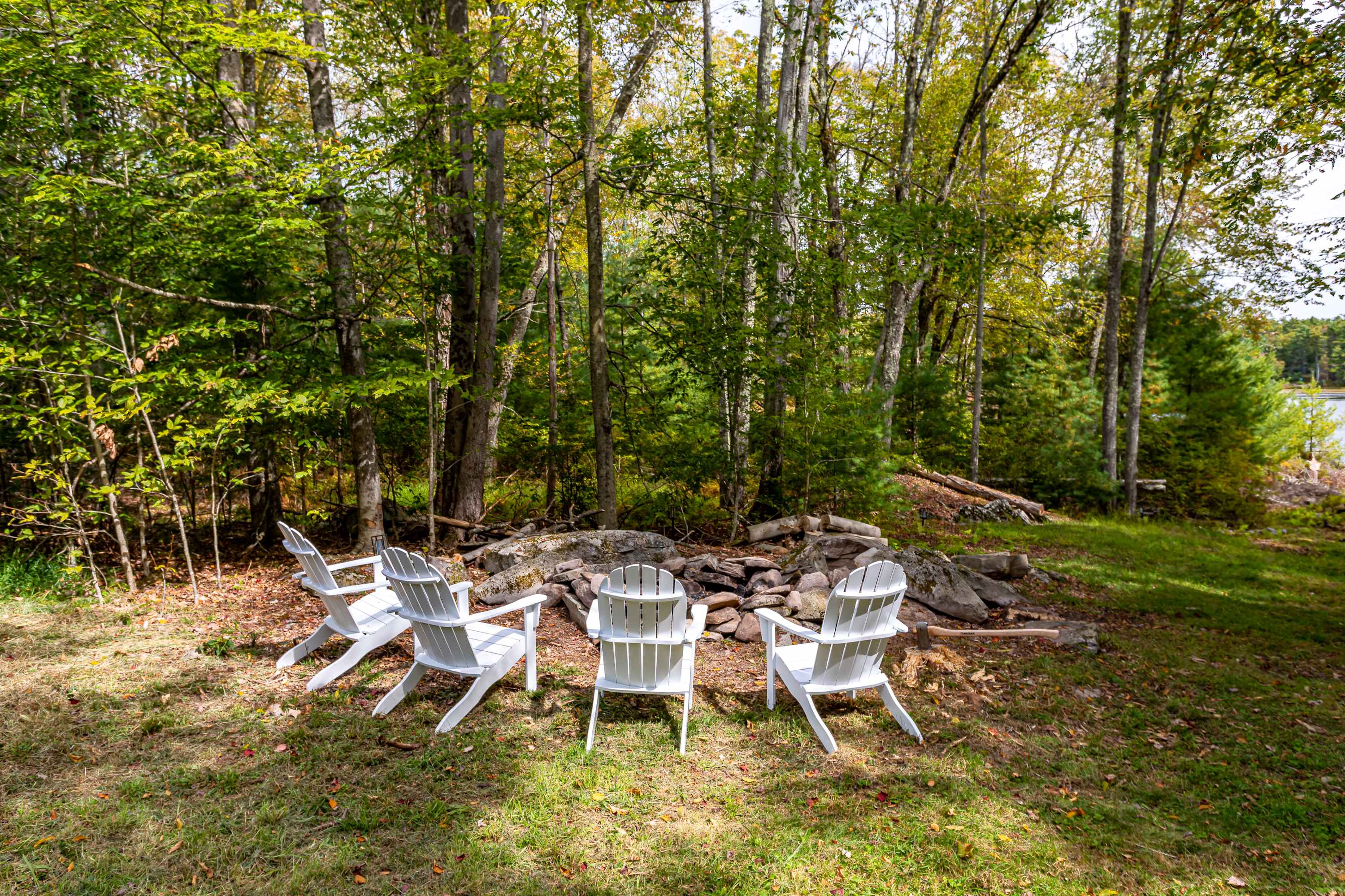 Outdoor fire pit area surrounded by four white Adirondack chairs, set on a grassy patch at the edge of a dense forest. A glimpse of a lake is visible through the trees in the background, creating a serene natural setting.