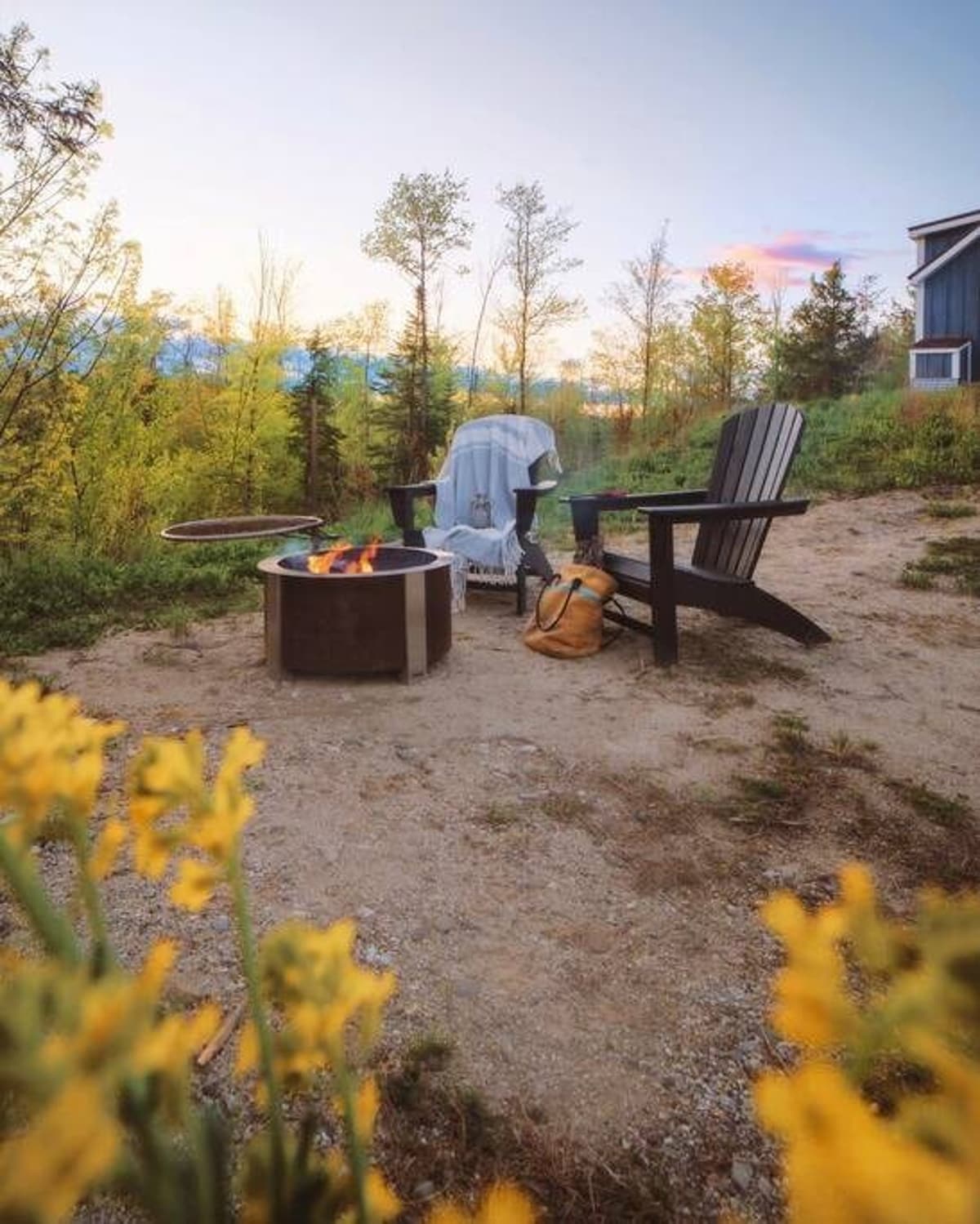 Outdoor fire pit area with a modern fire pit and two dark Adirondack chairs set on a gravel patch. Surrounded by lush greenery and trees under a soft sunset sky.