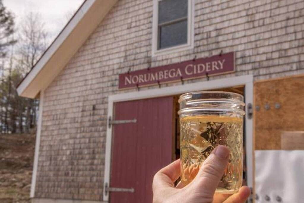 A hand holds a glass jar filled with a golden beverage, with a rustic building featuring a "NORUMBEGA CIDERY" sign and large red barn doors in the background. The building has light-colored shingle siding.