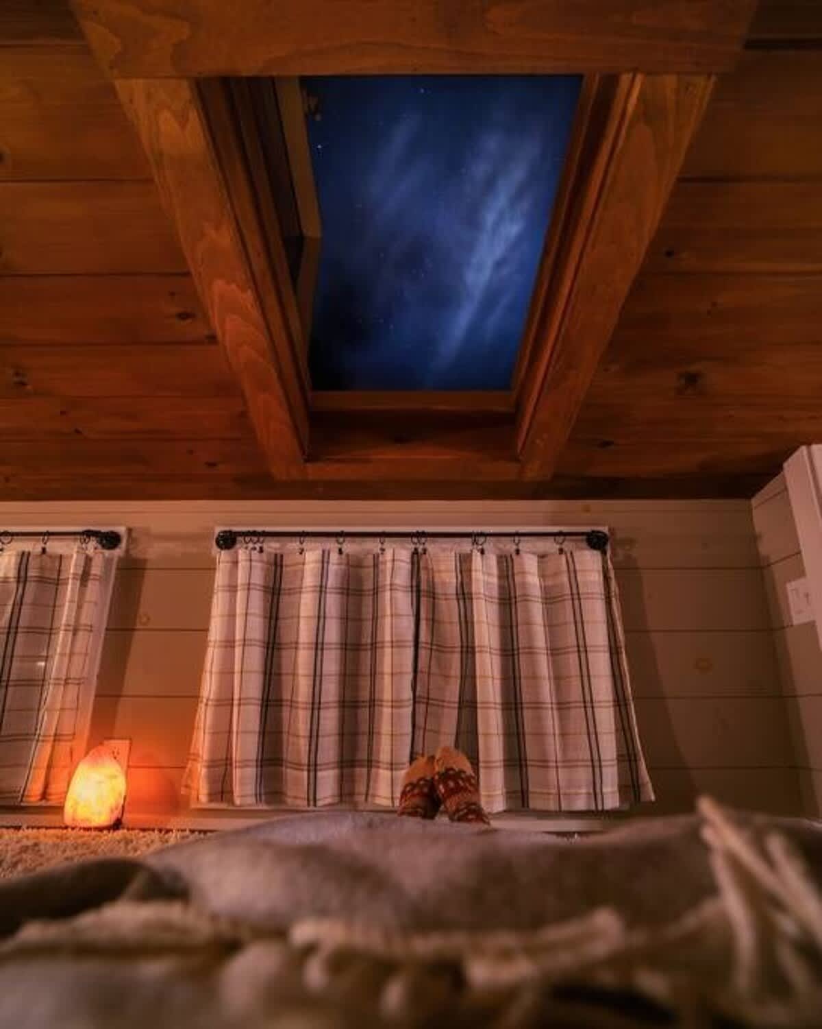 View from a cozy bed in a loft, looking up at a skylight revealing a night sky. The room features a wooden plank ceiling, windows with plaid curtains, and a glowing salt lamp.