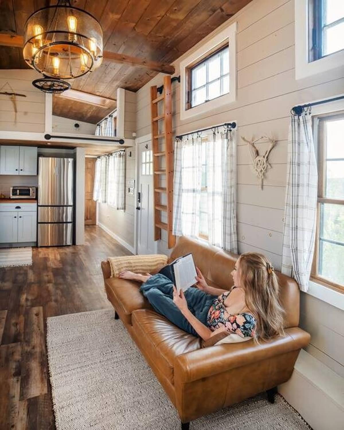 A cozy living area in a tiny house with a tan leather sofa, a light-colored area rug, and light gray shiplap walls. A wooden ladder leads to a loft, and a rustic chandelier hangs from the vaulted wooden ceiling.