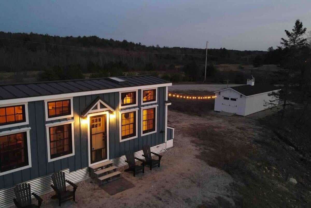 A tiny house with dark blue vertical siding and white trim, illuminated at dusk with warm interior lights and string lights. Two Adirondack chairs sit on a gravel patio in front of the entrance.