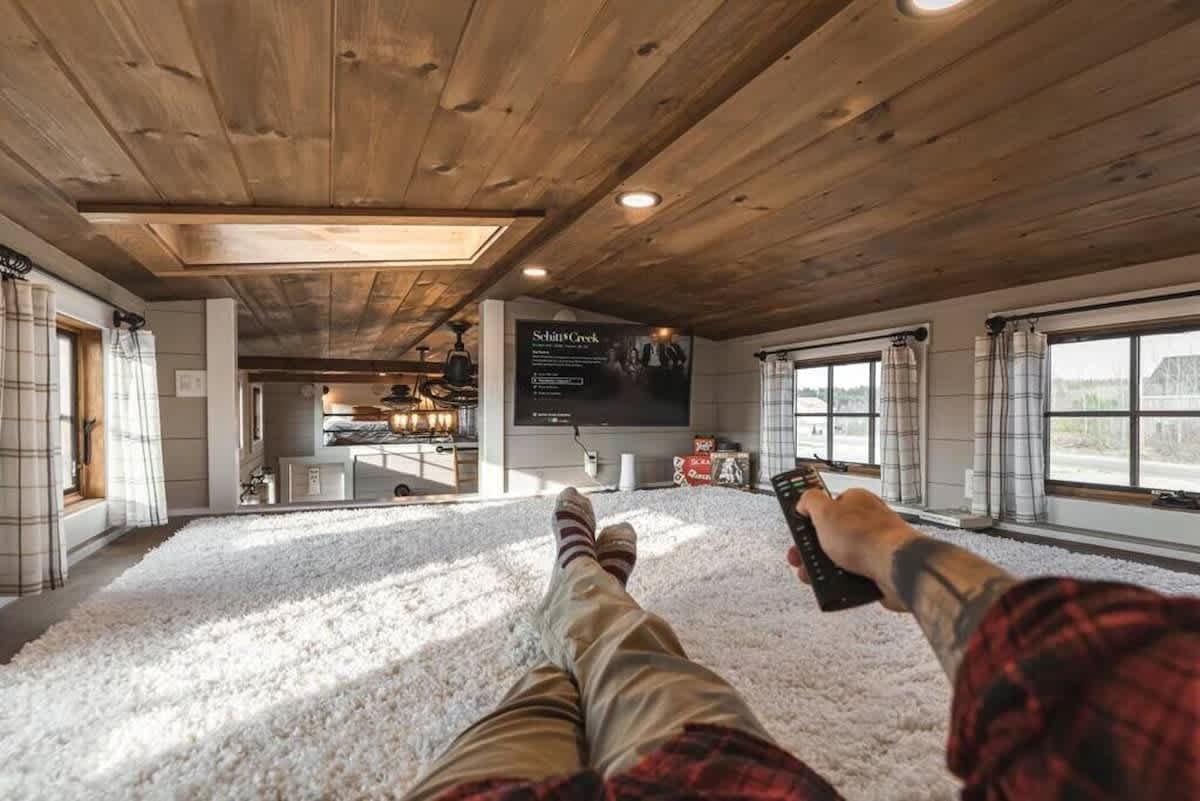 Elevated loft lounge area featuring a large wall-mounted TV and a plush white shag rug. Multiple windows with plaid curtains offer natural light, all beneath a warm wooden plank ceiling.