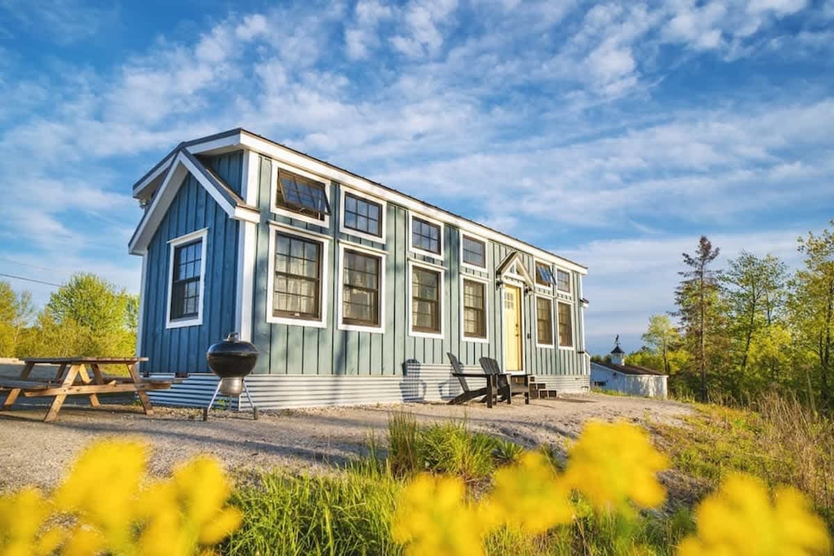 A long, narrow blue tiny house with white trim and numerous windows sits on a gravel patch under a bright blue sky. A picnic table and a BBQ grill are visible in the foreground, with yellow flowers blurring the bottom.