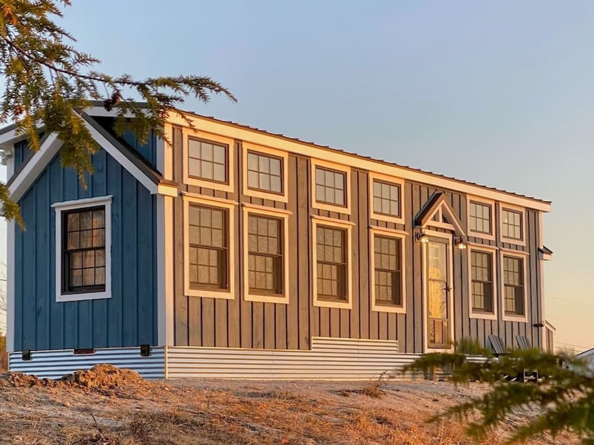 The tiny house exterior in daylight, showcasing dark blue vertical siding with white trim and numerous windows. A small porch roof covers the front door, and corrugated metal skirting is visible at the base.