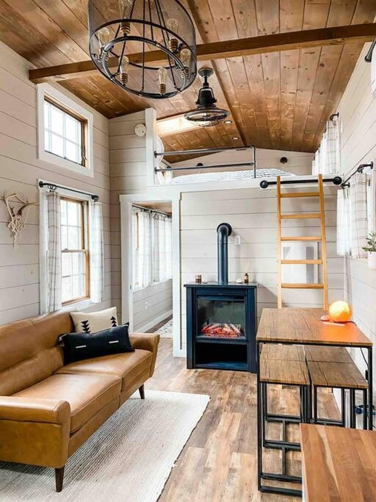 Cozy tiny house living room with a brown leather sofa, shiplap walls, and a wooden ceiling. A black electric fireplace sits below a loft bed accessible by a wooden ladder, with a small dining table nearby.