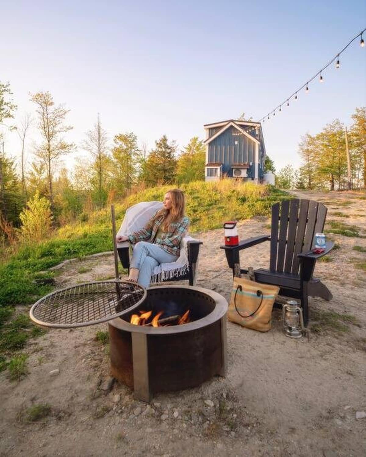 A woman relaxes in an Adirondack chair next to a metal fire pit with a grill, set on a dirt patch surrounded by trees. In the background, a blue tiny house sits on a hill under a clear sky.
