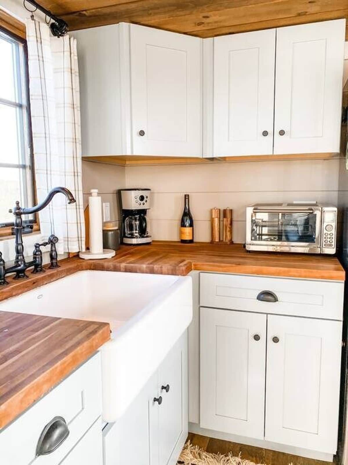 A cozy tiny house kitchen featuring light gray shaker cabinets, butcher block countertops, a white farmhouse sink with a black gooseneck faucet, and shiplap walls. Appliances include a coffee maker and a toaster oven.