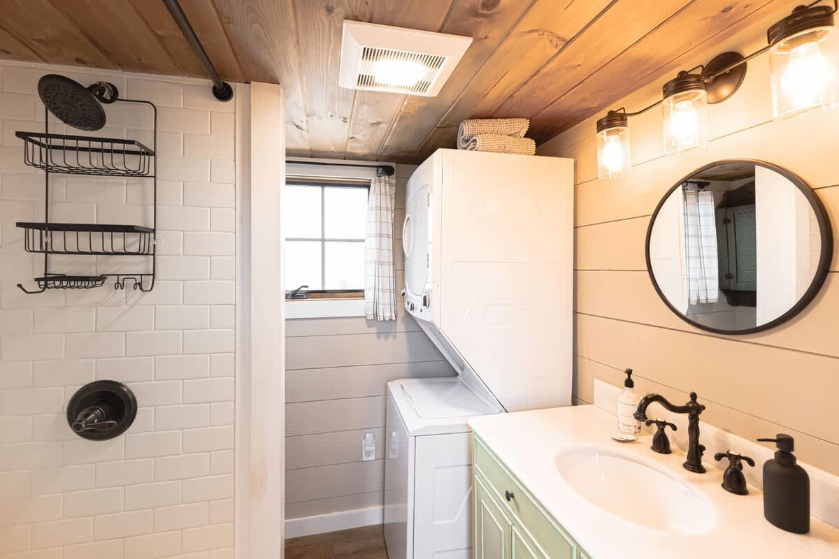 A modern bathroom features a white subway-tiled shower with black fixtures, a light green vanity with a white sink, and a stacked washer and dryer unit. The ceiling is wood-paneled, and a round mirror hangs above the sink.
