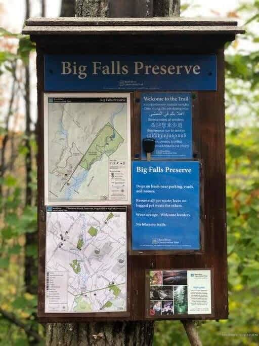 A wooden information board for "Big Falls Preserve" displays trail maps and rules, mounted on a tree trunk amidst lush green foliage. The sign includes details about pet policies and trail etiquette.