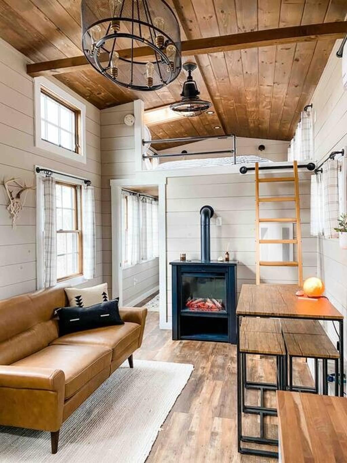 Cozy tiny house living room with a brown leather sofa, shiplap walls, and a wooden ceiling. A black electric fireplace sits below a loft bed accessible by a wooden ladder, with a small dining table nearby.