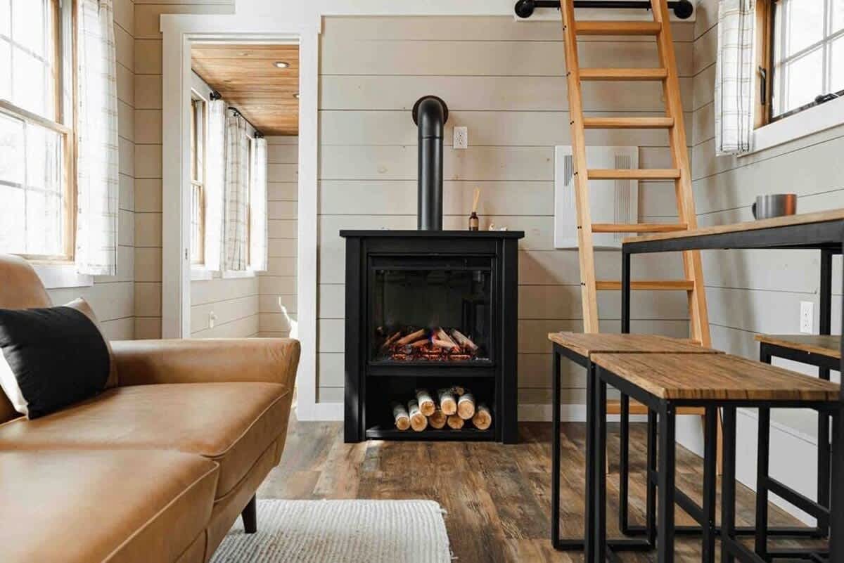 A warm living space featuring a black electric fireplace with a stovepipe, flanked by a tan leather sofa and a small dining table with two stools. Light gray shiplap walls and a wooden ladder to a loft are visible.