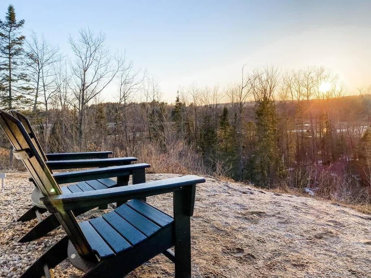 Two dark Adirondack chairs positioned on a gravelly slope, overlooking a vast natural landscape of trees and hills. The scene is bathed in the warm light of a sunrise or sunset.