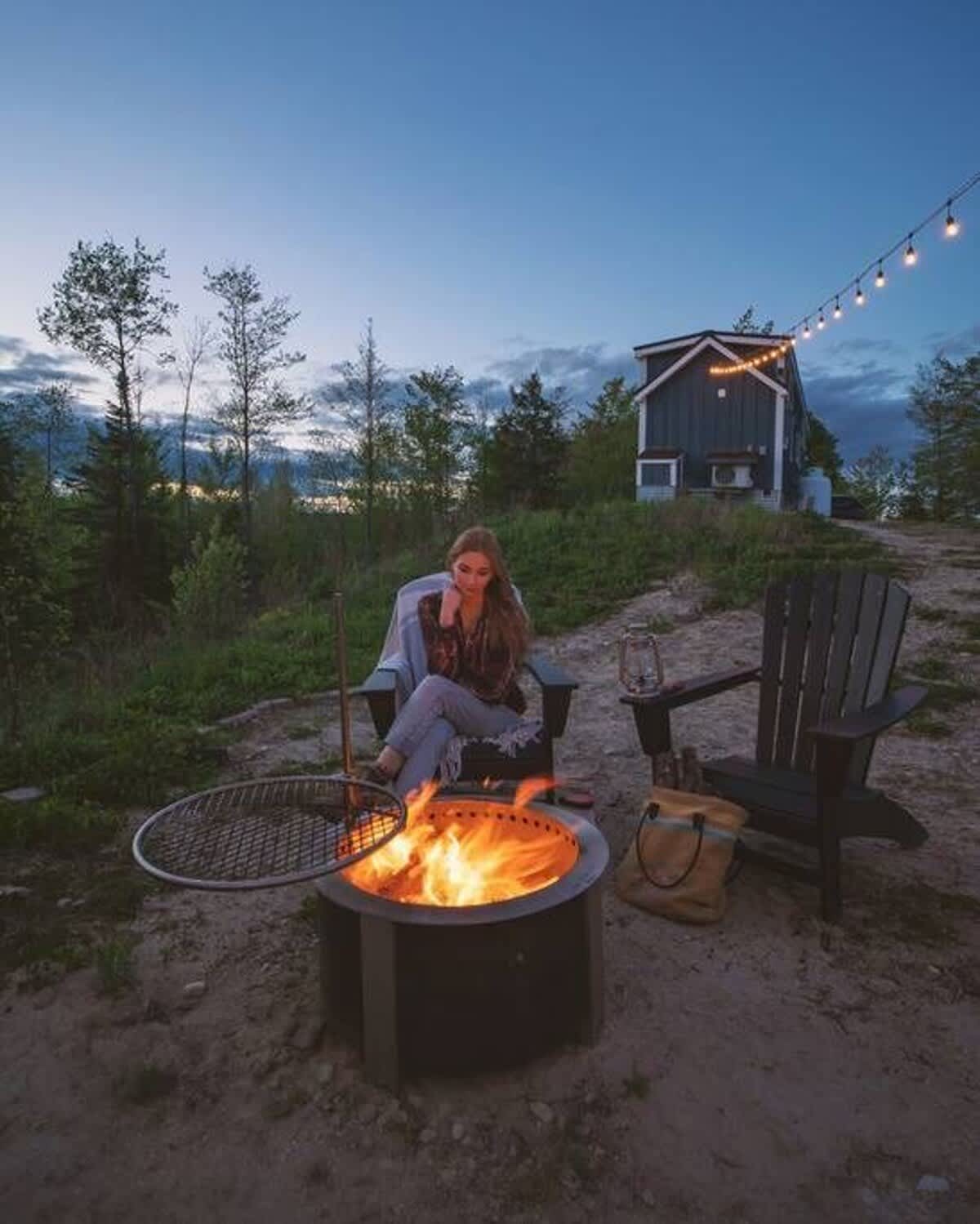 A woman sitting in an Adirondack chair by a metal fire pit with a grill, enjoying a fire at dusk. The tiny house is visible in the background, with string lights illuminating the outdoor area.