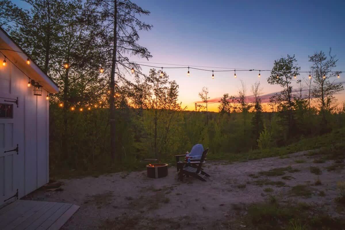 Evening view of an outdoor fire pit area with string lights illuminating a small white cabin and the surrounding trees. A single Adirondack chair faces the glowing fire pit under a colorful sunset sky.