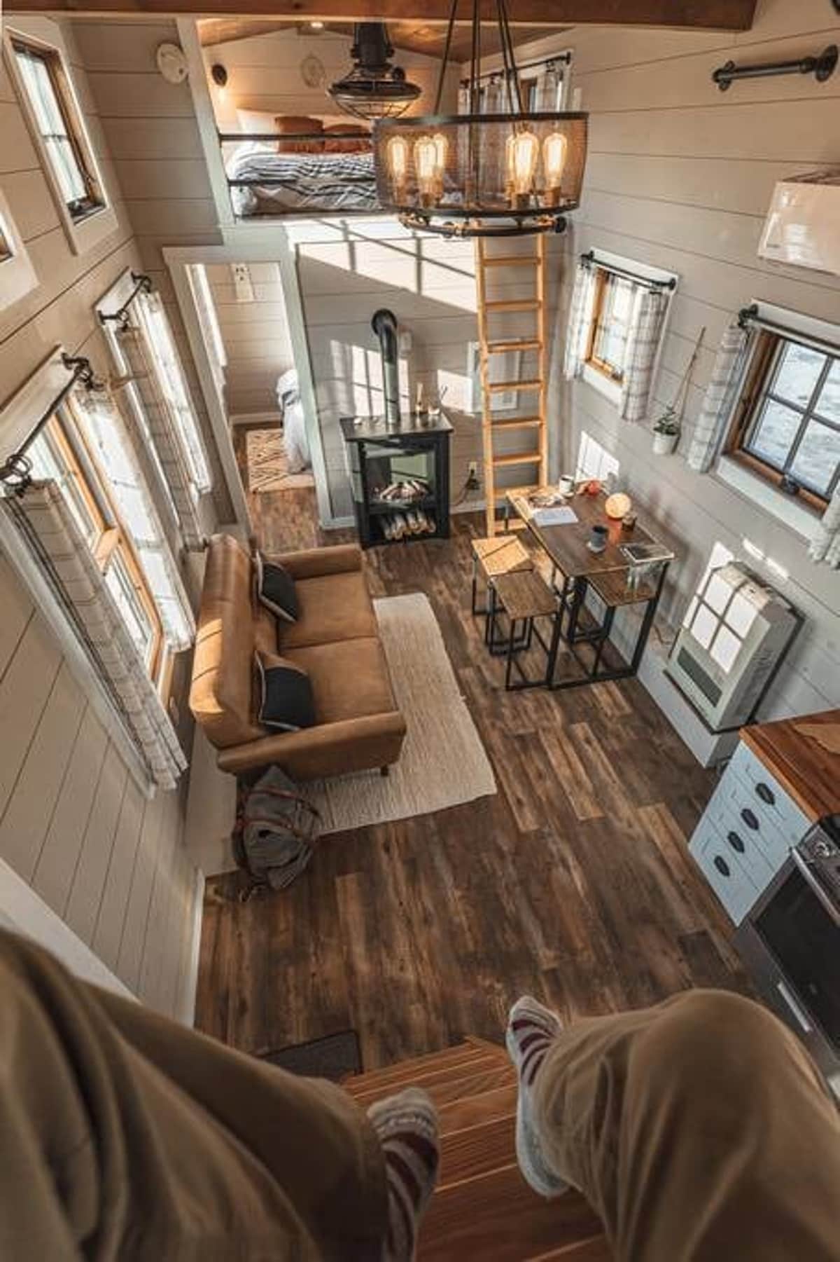 High-angle view of a tiny house interior featuring a brown sofa, a small dining table, a wood-burning stove, and a ladder leading to a loft bedroom. The space has light wood-paneled walls and dark wood flooring.