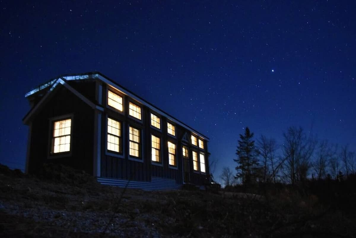 A long shot of the tiny house at night, with all interior lights glowing warmly against a dark, starry sky. The house features multiple windows and dark siding, nestled in a natural landscape.