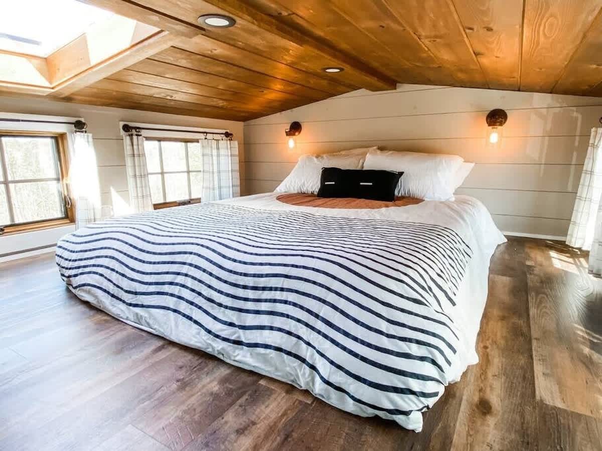 A bright loft bedroom with a large bed featuring a black and white striped duvet, set under a vaulted wooden ceiling with skylights. Light gray shiplap walls and warm sconce lighting complete the rustic modern look.