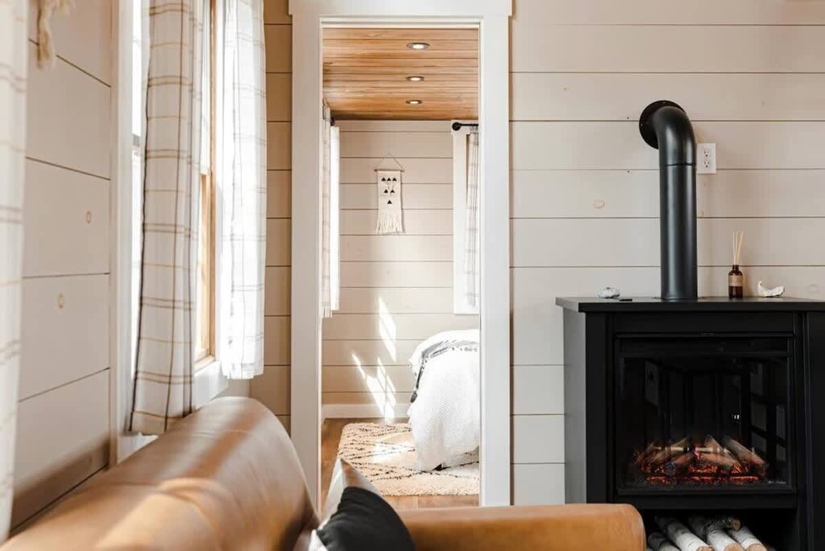 Cozy living area with a light brown sofa and a black electric fireplace, featuring light shiplap walls and wood plank flooring. A doorway leads to a bright bedroom with a bed and a rug.