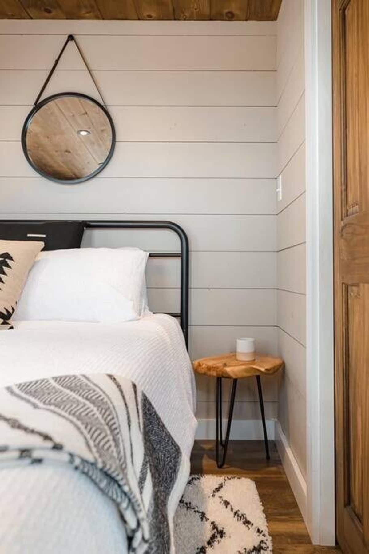 A rustic bedroom with light gray shiplap walls, a black metal bed frame with white bedding, and a small live-edge wood side table. A round black-framed mirror hangs above the bed.