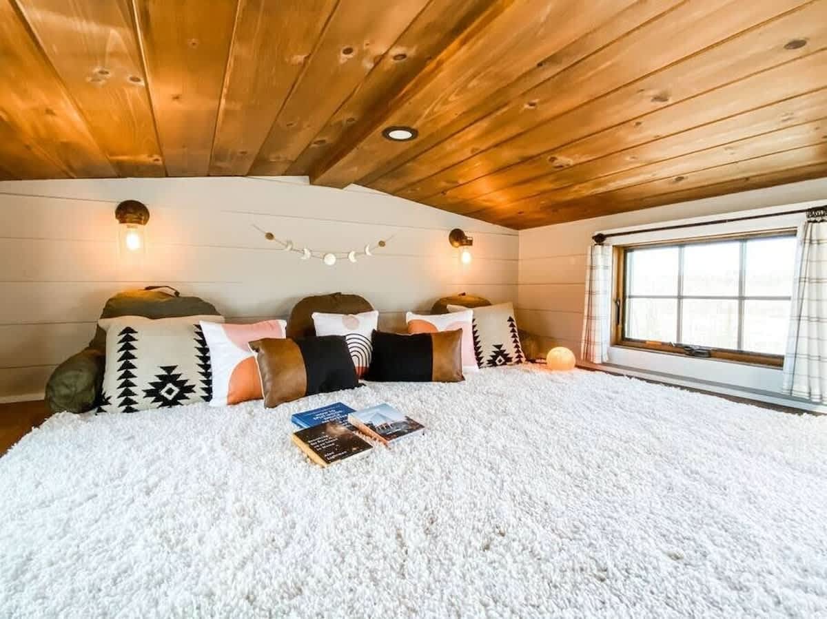 Spacious loft bedroom with a plush white shag rug covering the floor, adorned with an assortment of decorative pillows. Wooden plank ceiling, wall sconces, and a window with plaid curtains complete the cozy space.