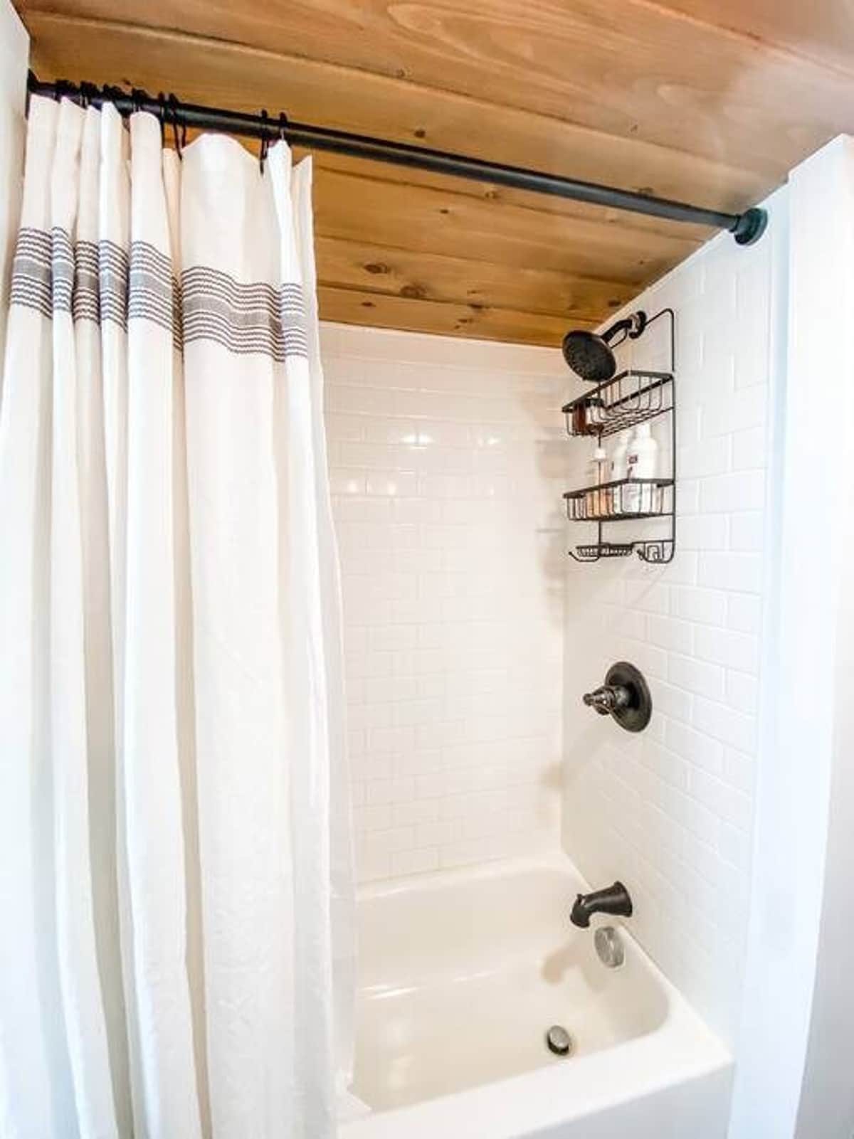 Bright bathroom featuring a white subway tile shower with a tub, black fixtures, and a white striped shower curtain. A black shower caddy holds toiletries, and the ceiling is wood-paneled.