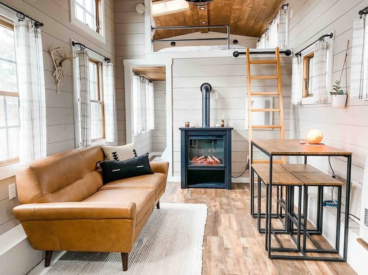 Spacious living area with a light brown sofa, a black electric fireplace, and a dining table with stools. A ladder leads to a loft, and the kitchen with light blue cabinets and stainless steel appliances is visible on the right.