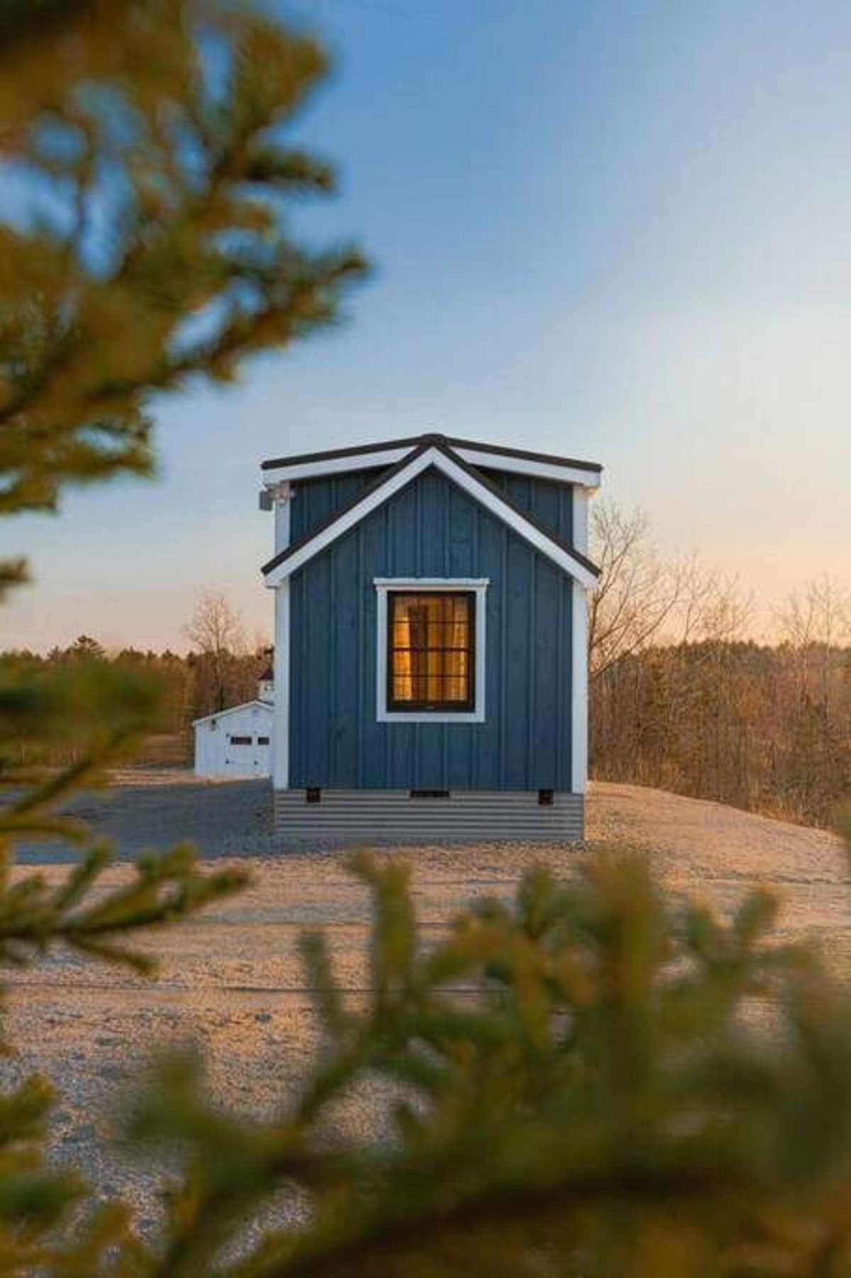 A small, blue tiny house with a dark roof and white trim stands on a gravel lot at sunset, framed by blurred green pine branches in the foreground. A warm light glows from its single visible window.
