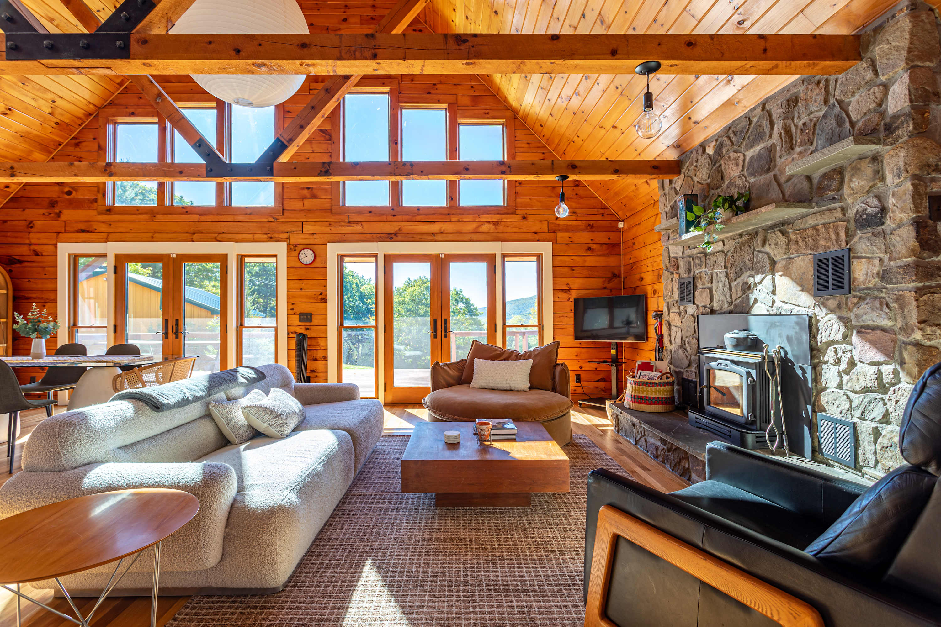 Spacious living room with vaulted wood-paneled ceilings and walls, featuring a large stone fireplace, a light-colored sectional sofa, and multiple windows offering bright natural light and views.