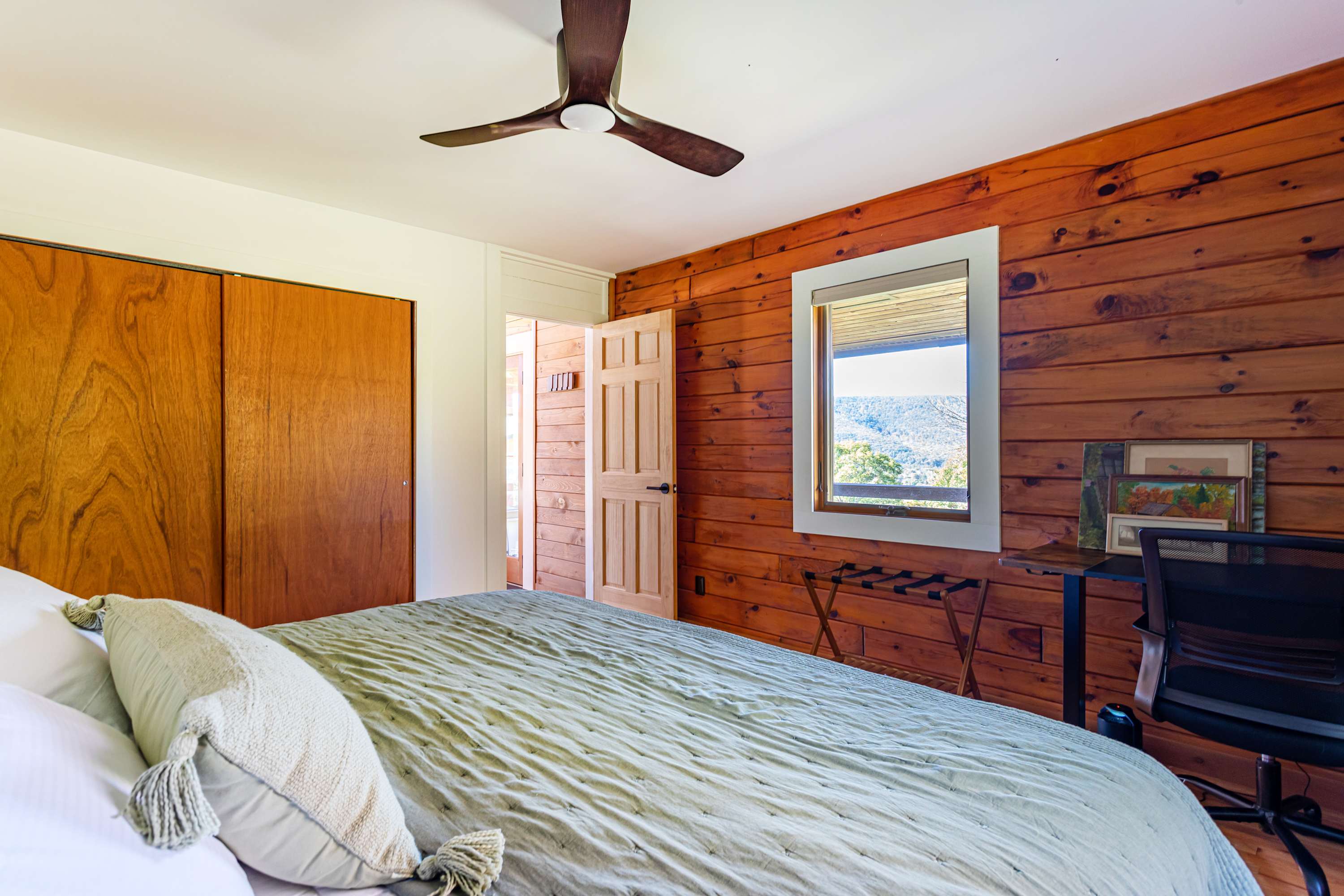 Another angle of bedroom-3 showing the queen bed with a green throw, a wood-paneled wall, and a closet with wooden bifold doors. A window offers views of the outdoors.