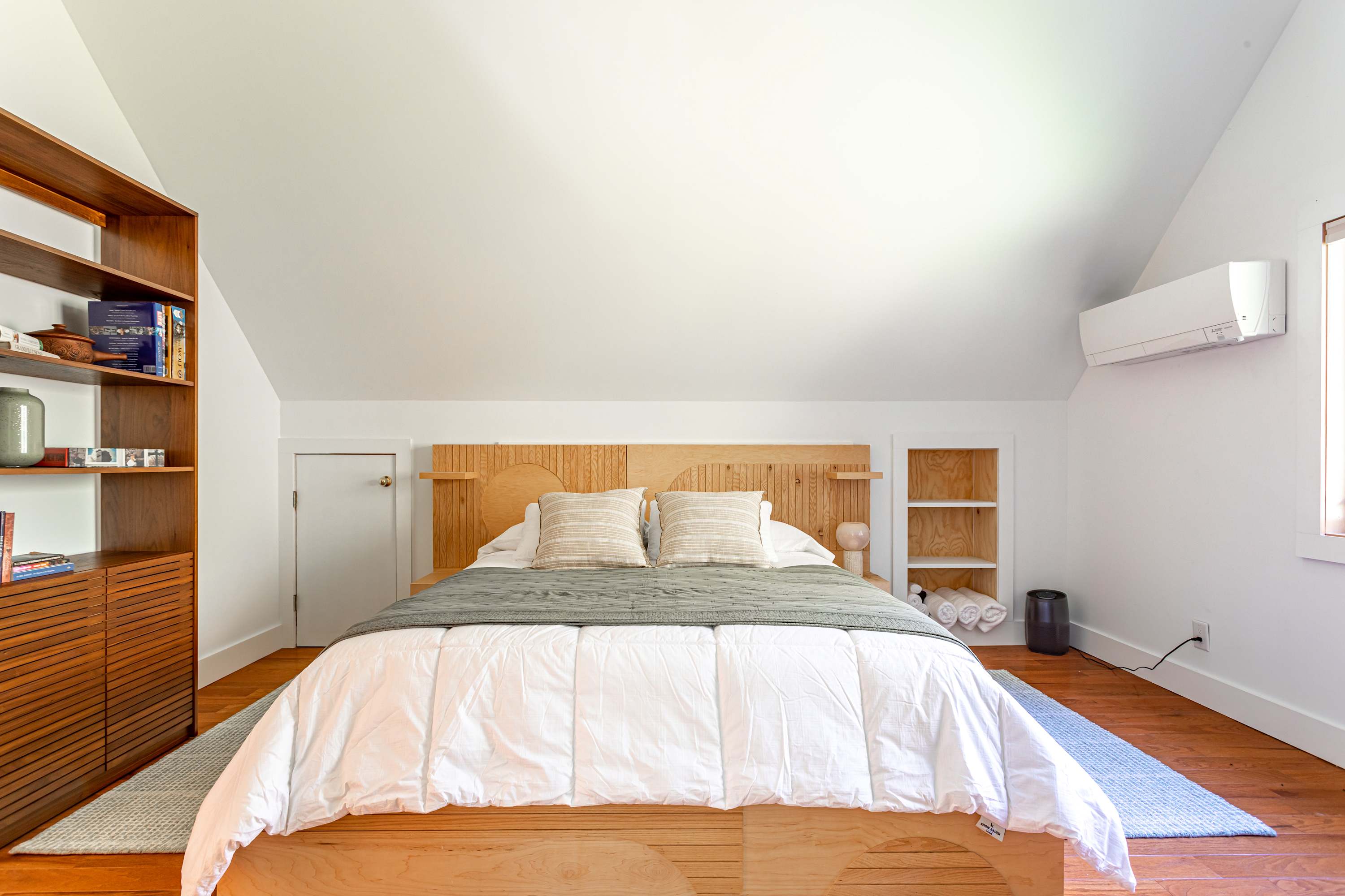 Bright bedroom-1 with a light wood platform bed and headboard, white walls, and a sloped ceiling. Built-in shelving and a modern air conditioning unit are visible.