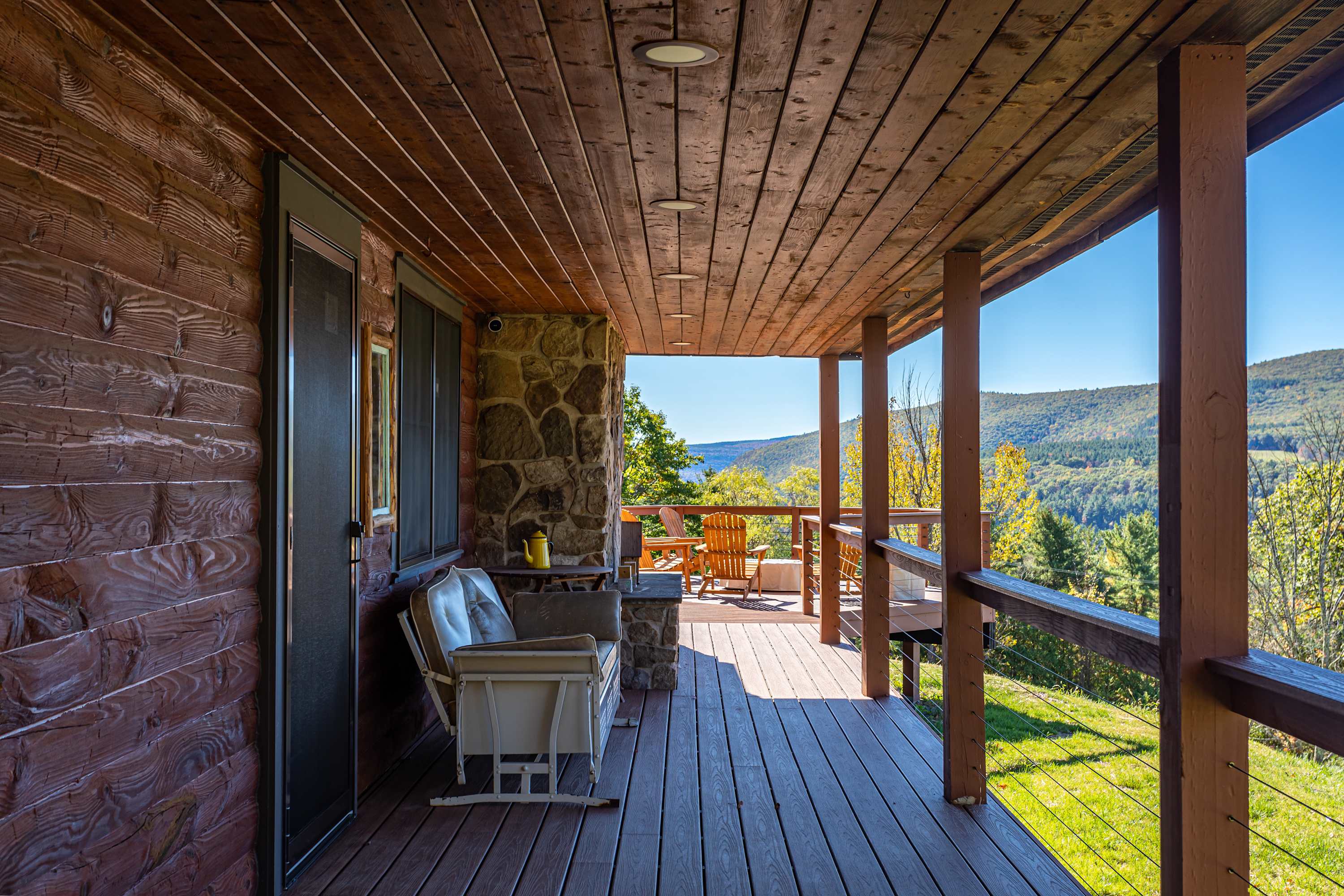 Covered wooden porch with a rocking chair and stone accents on the cabin wall, offering glimpses of distant mountains and a sunny outdoor deck.