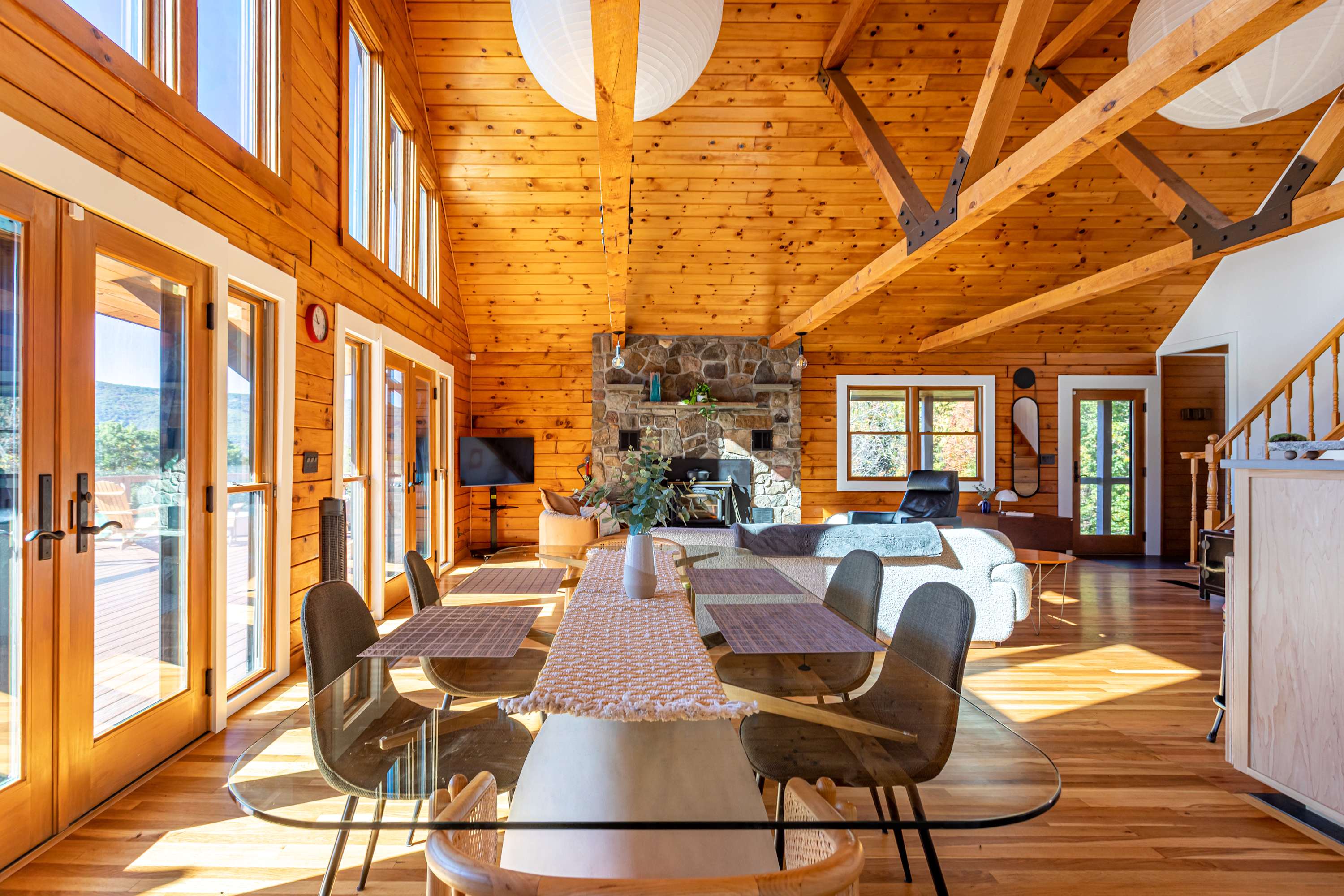 Spacious open-concept dining and living area with a glass dining table, leading to a comfortable living space centered around a large stone fireplace, all under a high vaulted wooden ceiling with exposed beams.